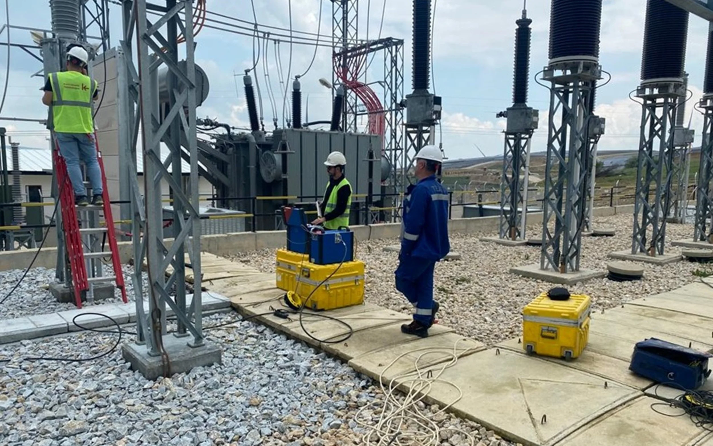 Workers in safety gear performing maintenance or inspection at a power substation with electrical infrastructure and equipment.