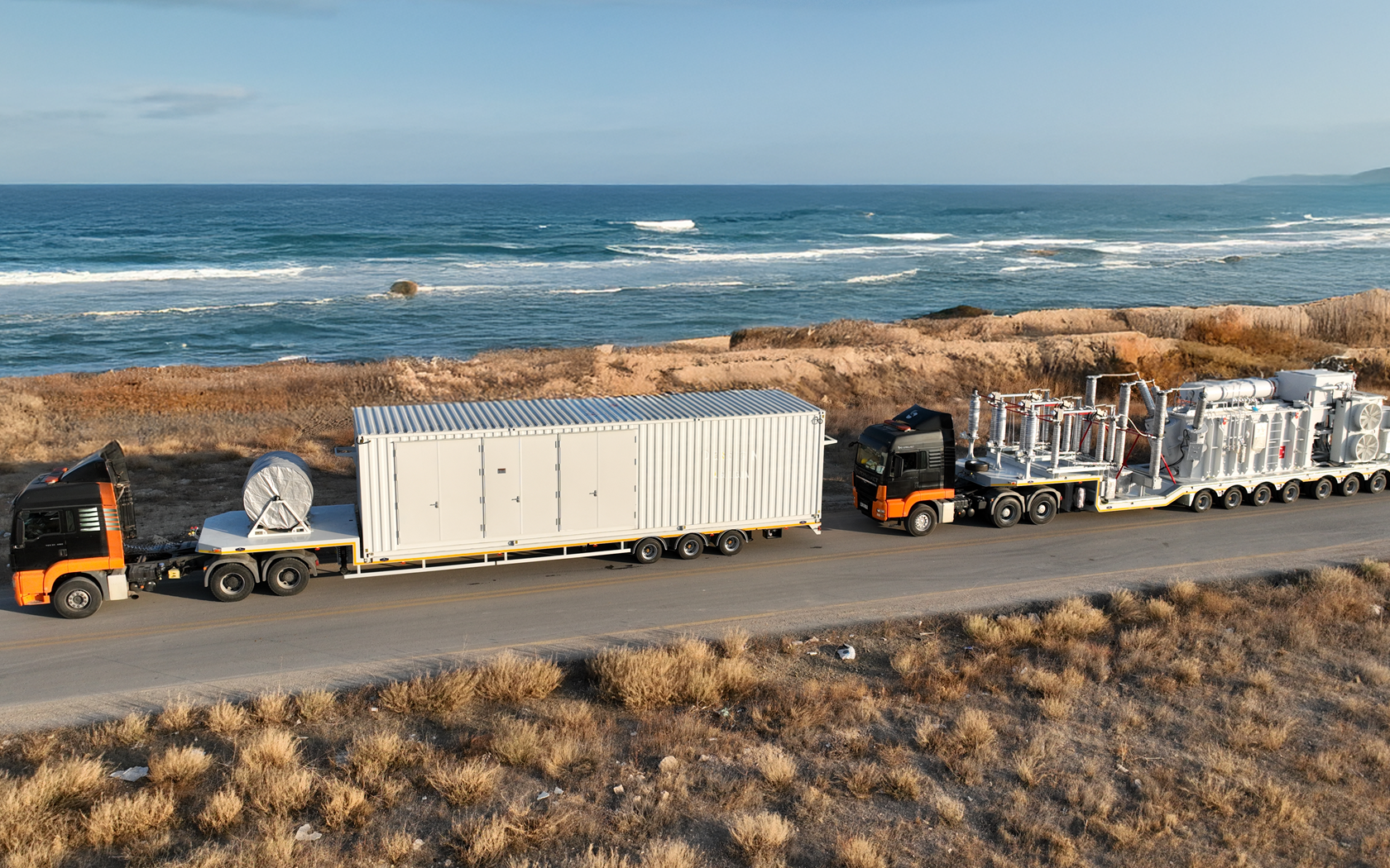 A large black semi-truck with an orange stripe on its cab transports a container and a satellite dish, and is traveling along a coastal road near the ocean with rocky and grassy terrain on either side.
