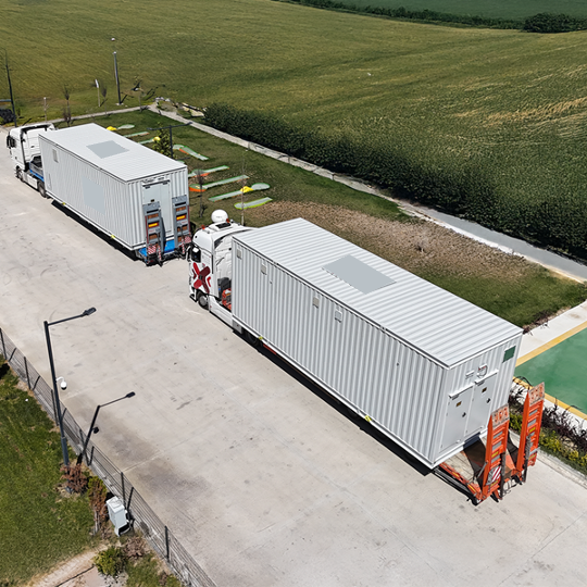 Two large white shipping containers on flatbed trucks parked on a paved road near a grassy field and a playground area.