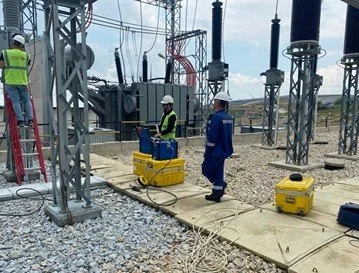 Electric utility workers wearing safety gear working at a power substation with equipment and tools, including yellow toolboxes and a ladder.