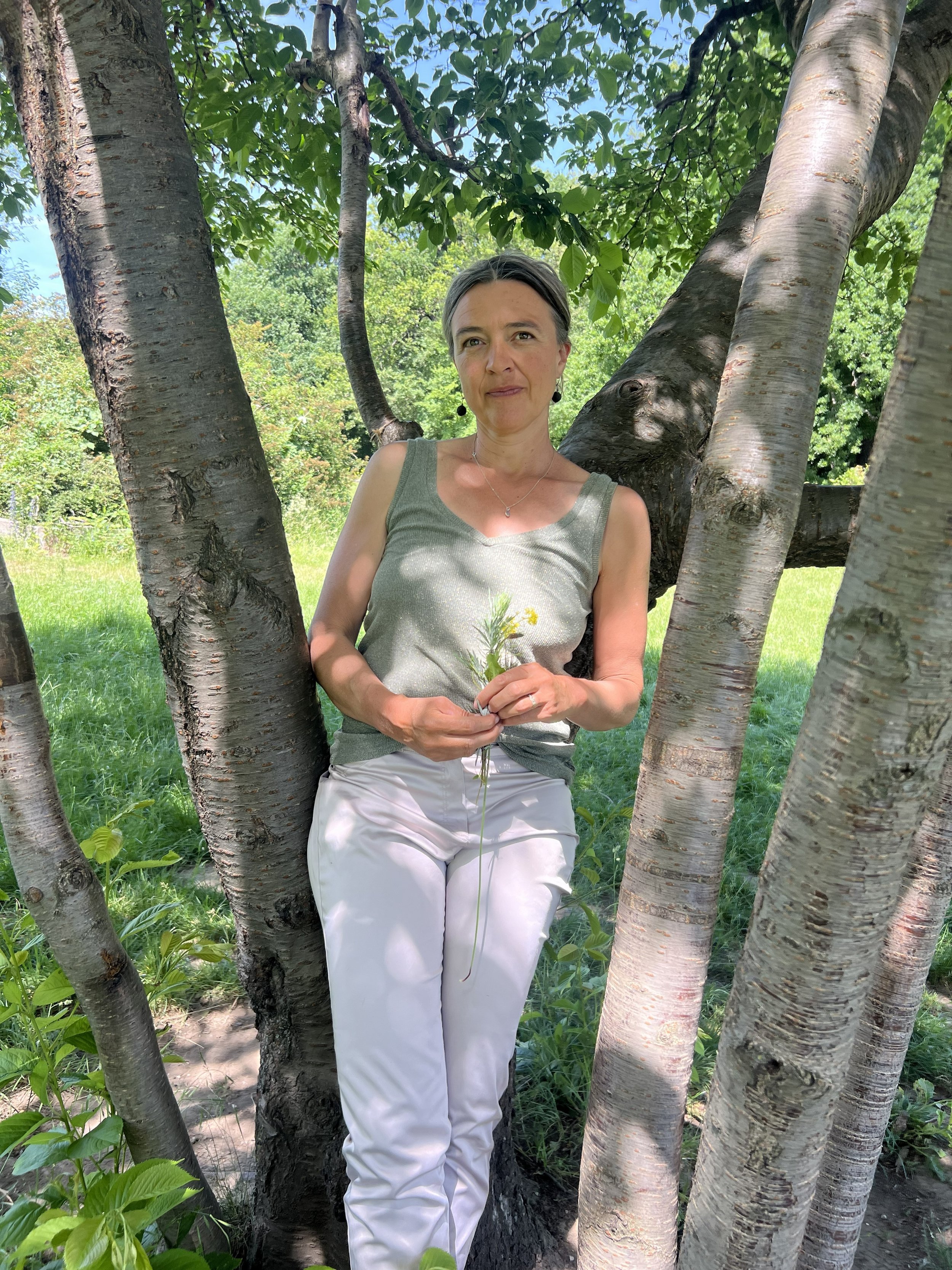 Eine Frau steht zwischen mehreren Bäumen mit grünen Blättern im Freien an einem sonnigen Tag, hält eine kleine Blumenstrauß in den Händen und sieht in die Kamera.