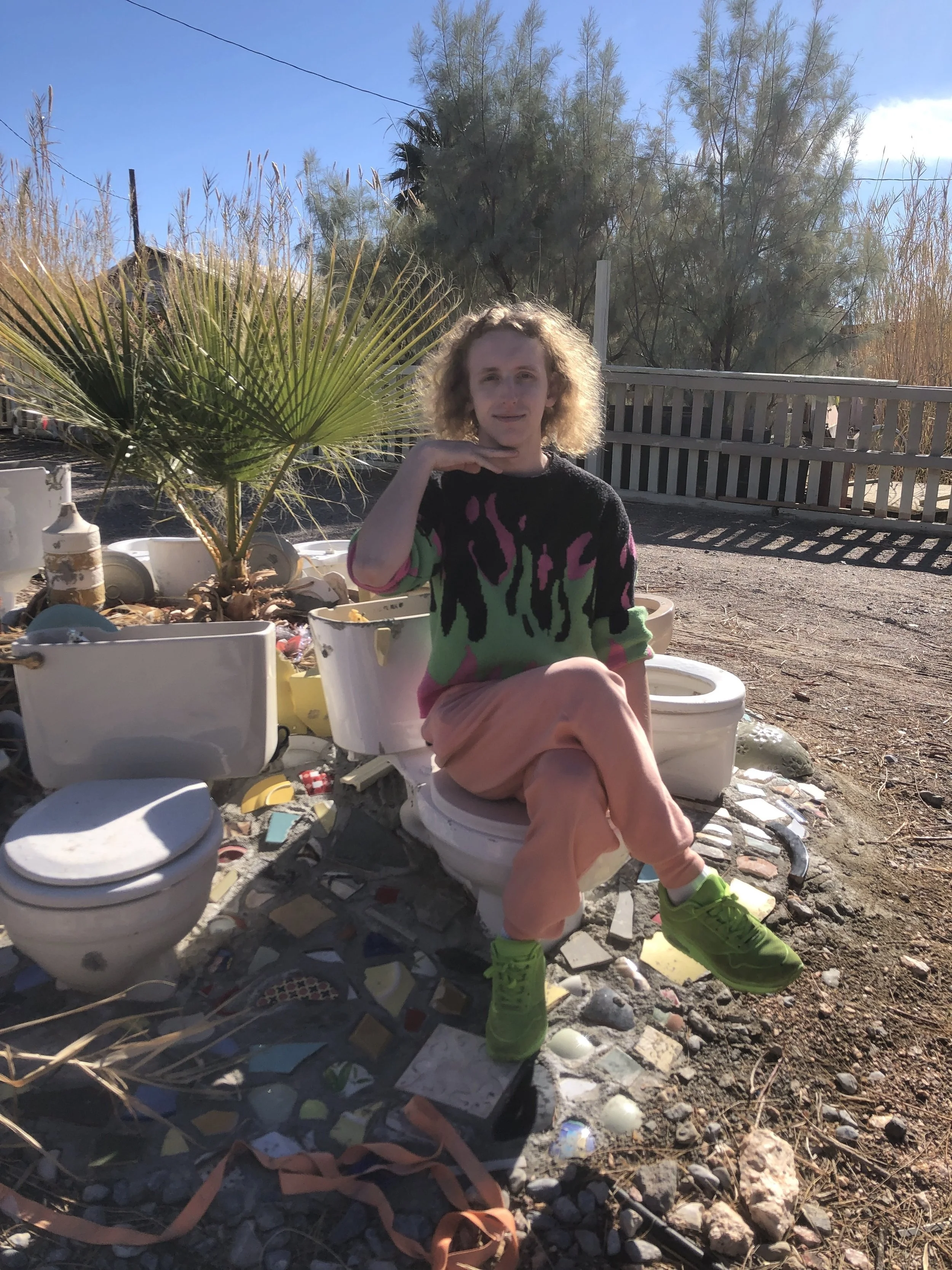 A blonde nonbinary person in colorful clothes posing while sitting on a sculpture made of several empty toilets arranged in a circle.