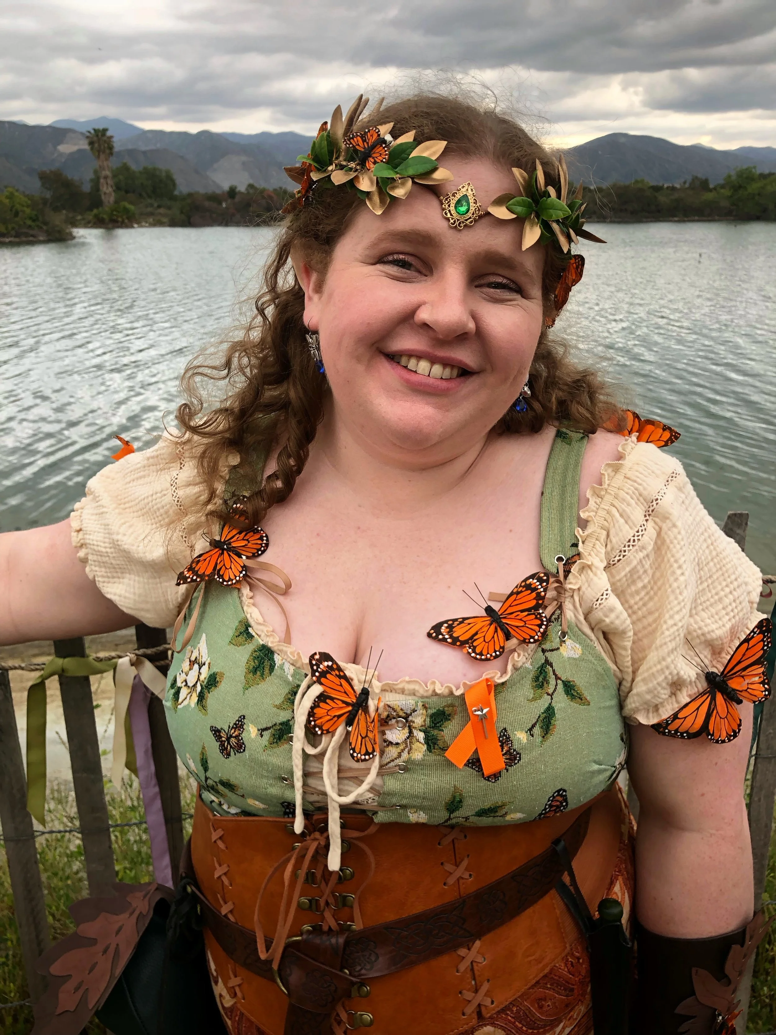 A woman dressed in fairy or elf costume with monarch butterflies attached to her outfit and headpiece, standing near a body of water with mountains and cloudy sky in the background.