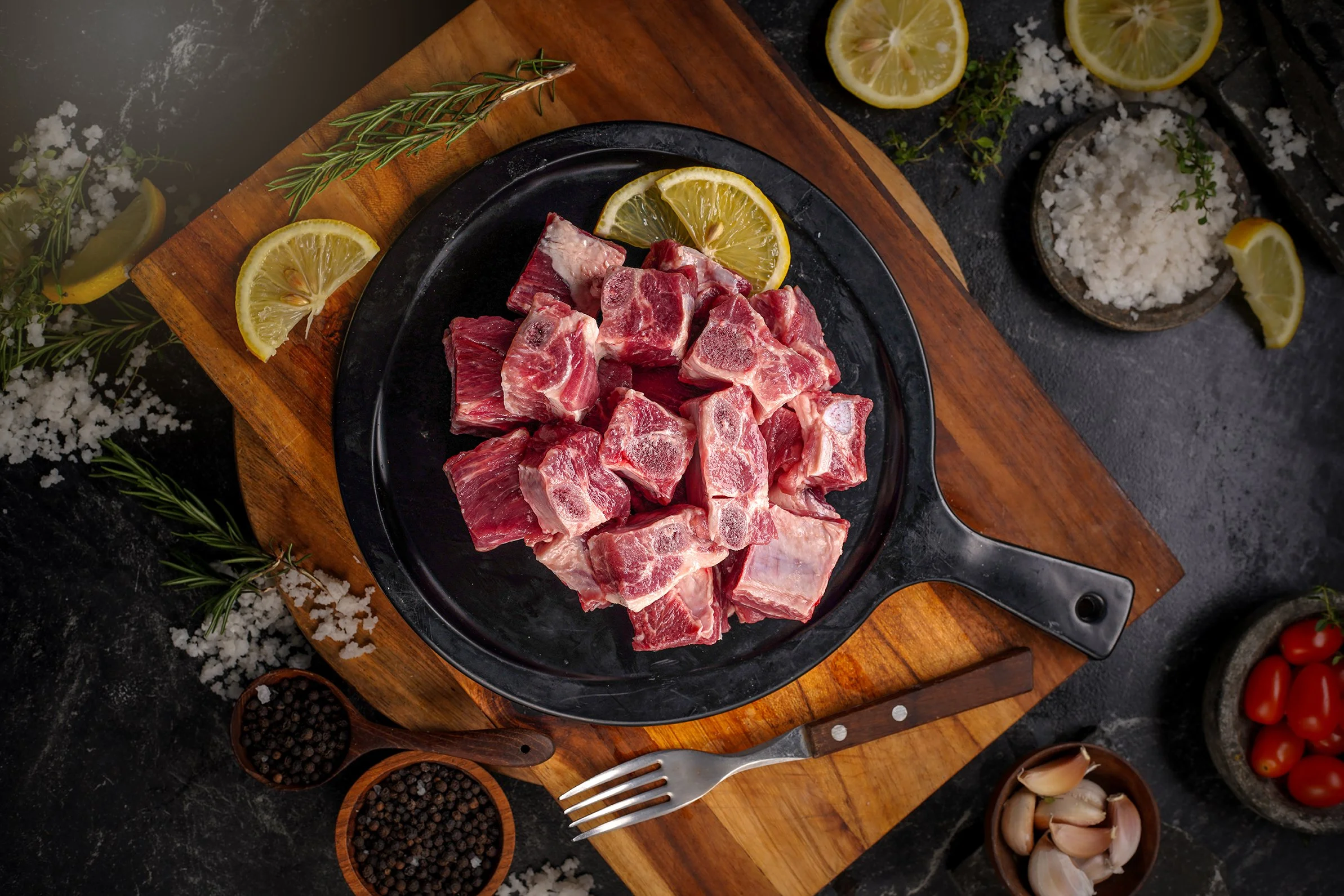 Raw beef pieces on a black plate with lemon slices, surrounded by herbs, garlic, cherry tomatoes, and salt on a dark countertop.