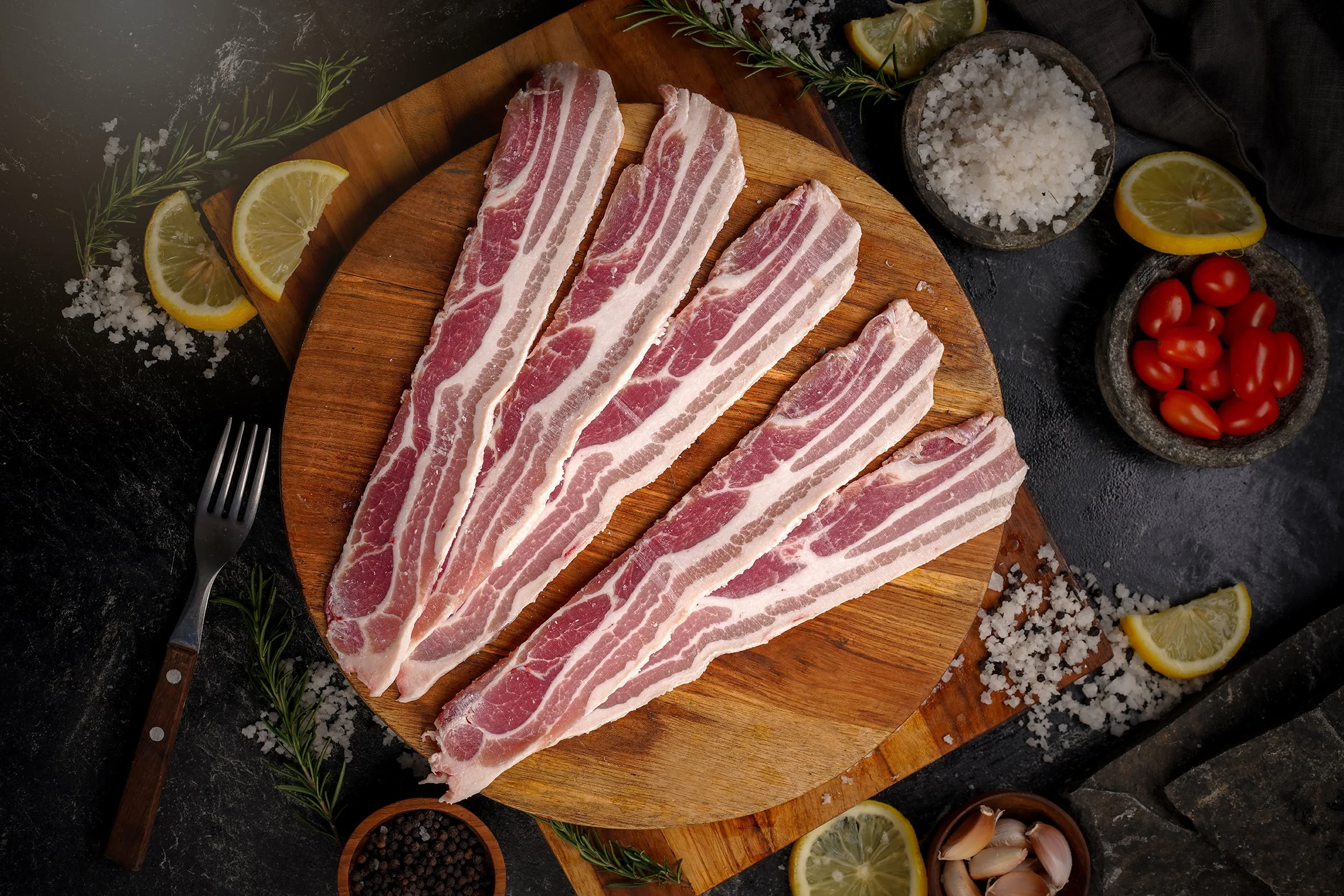 Raw bacon slices on a wooden cutting board surrounded by lemon slices, cherry tomatoes, garlic, salt, black pepper, rosemary, and other seasonings.