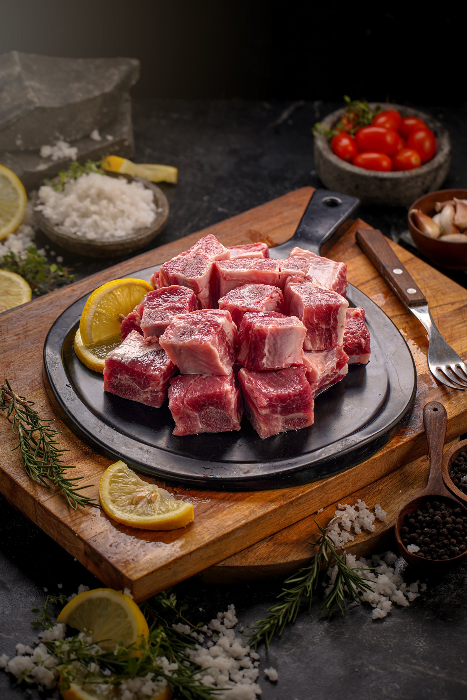Raw beef short ribs on a black plate surrounded by lemon slices, herbs, garlic, cherry tomatoes, and salt on a wooden cutting board.