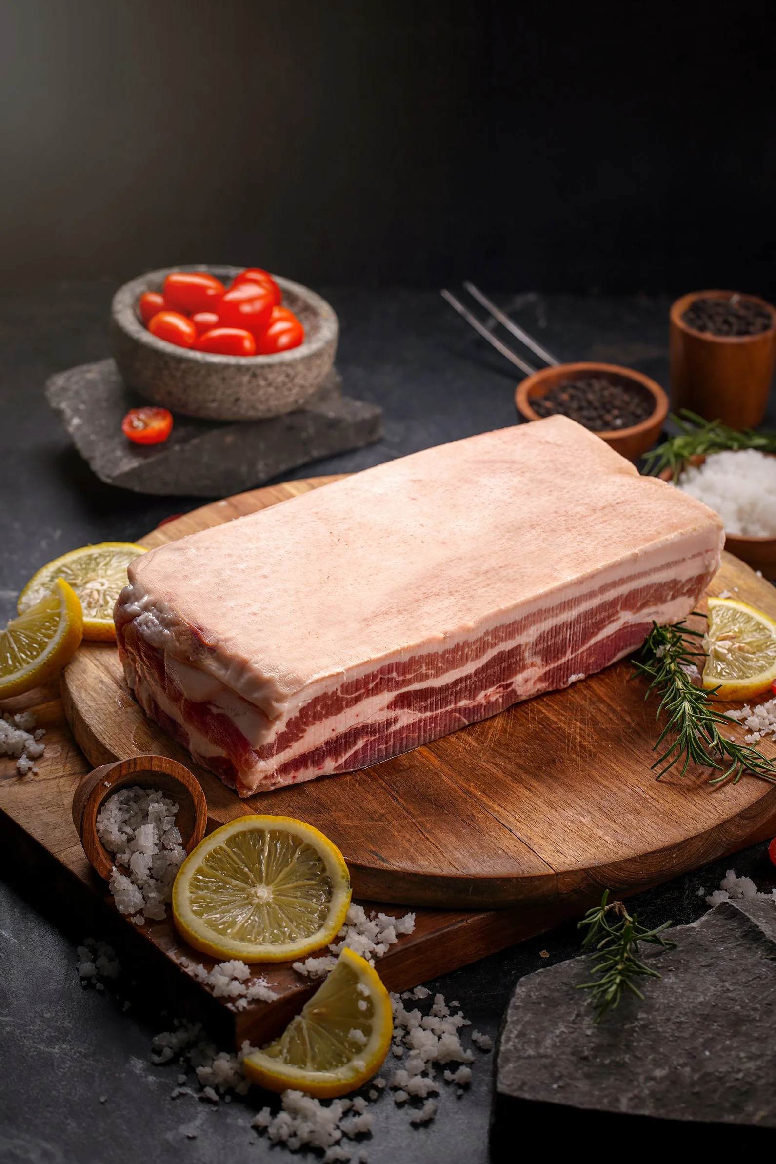 A raw strip of pork belly on a wooden cutting board, garnished with lemon slices and fresh herbs, with cherry tomatoes, lemon slices, salt, and pepper in the background.