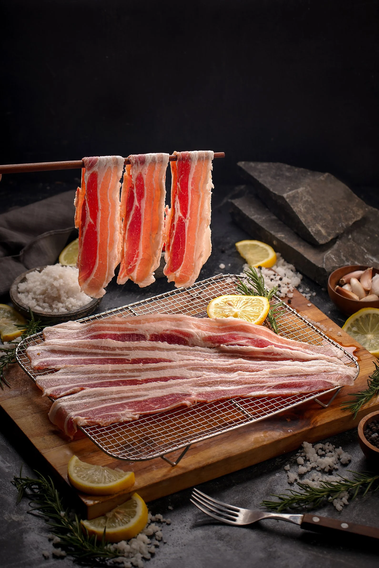 Raw bacon slices hanging on a wooden rod, with slices of lemon, rosemary, coarse salt, and mushrooms on a dark background.