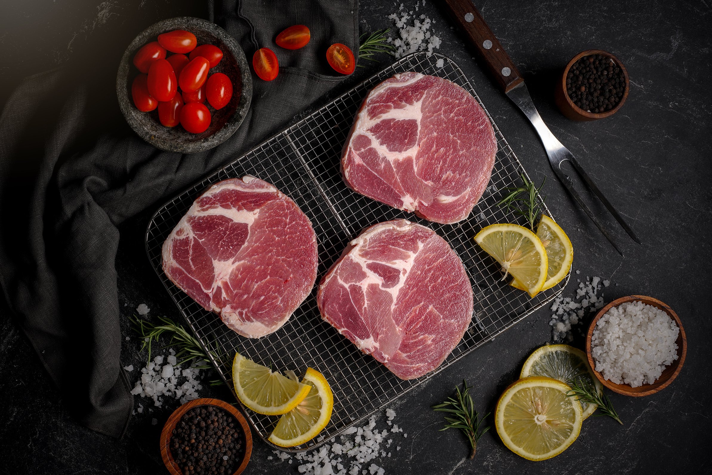 Raw pork chops on a wire rack with lemon slices, cherry tomatoes, salt, black pepper, and herbs on a dark surface.