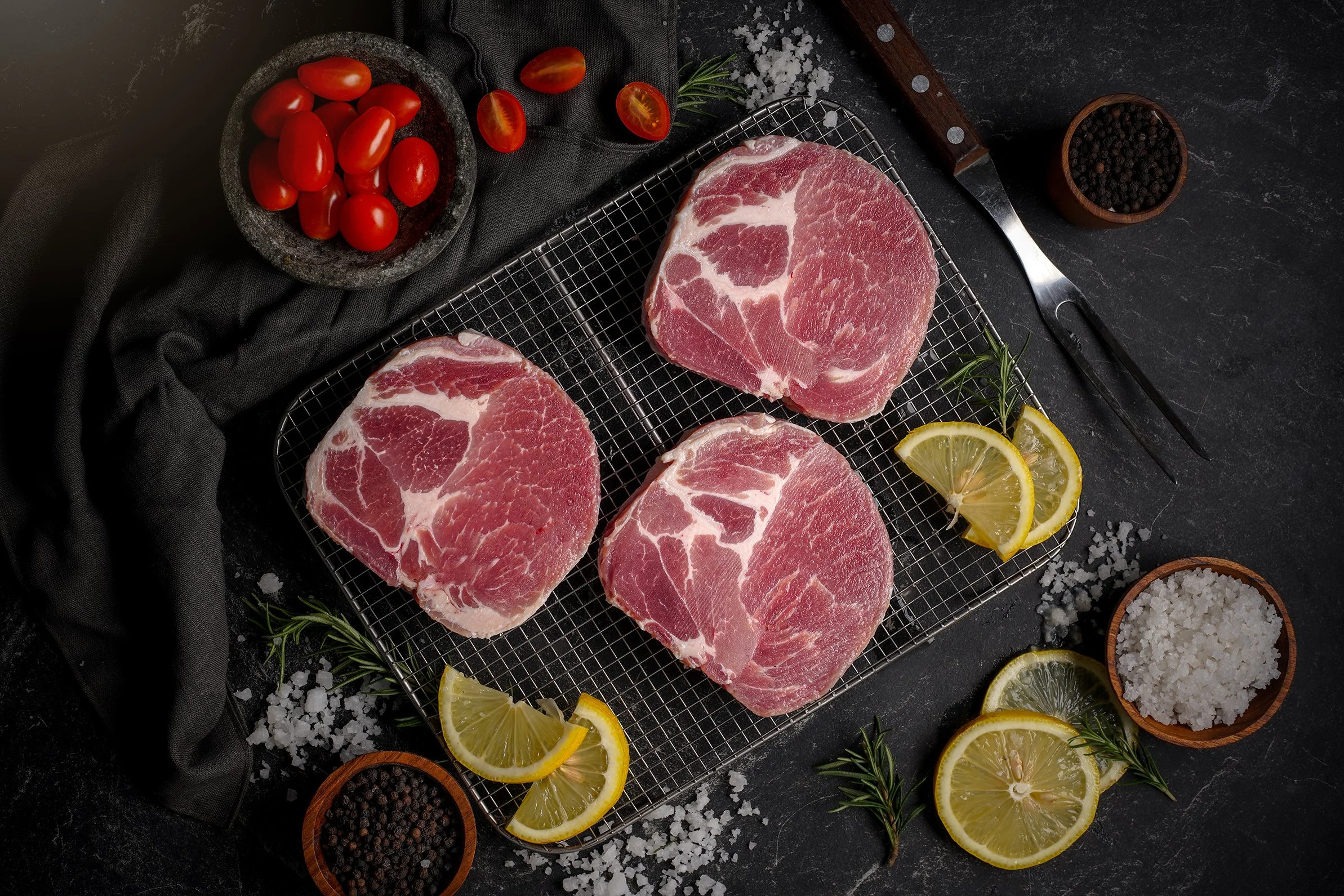 Four raw pork chops on a metal cooling rack, surrounded by sliced lemons, black pepper, cherry tomatoes, and salt, with herbs and salt scattered around on a dark surface.