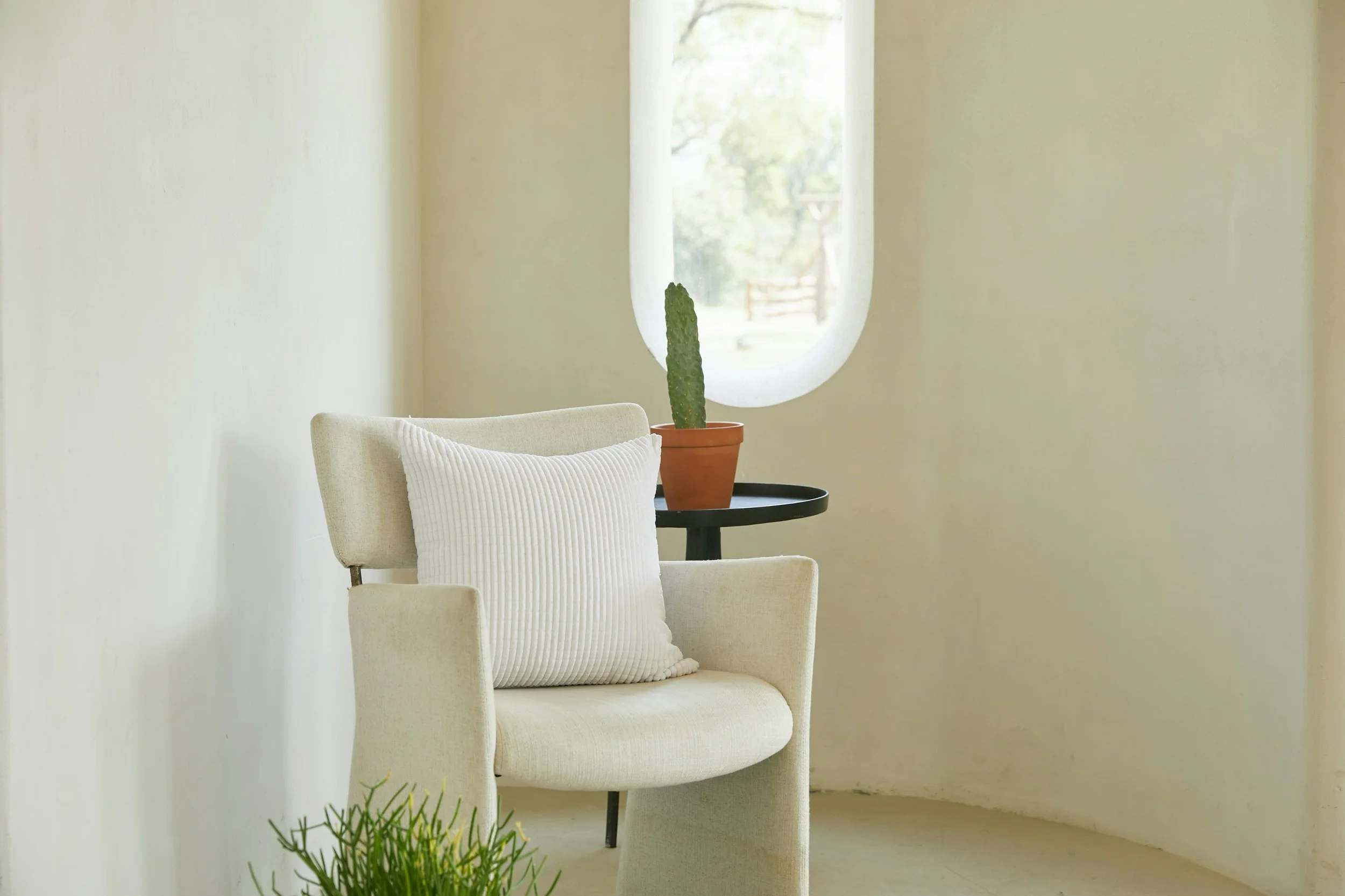 Un fauteuil beige avec un coussin blanc à rayures, à côté d'une petite table ronde noire avec une plante cactus en pot. En arrière-plan, un mur avec une fenêtre laissant passer la lumière. Le décor est minimaliste et lumineux.