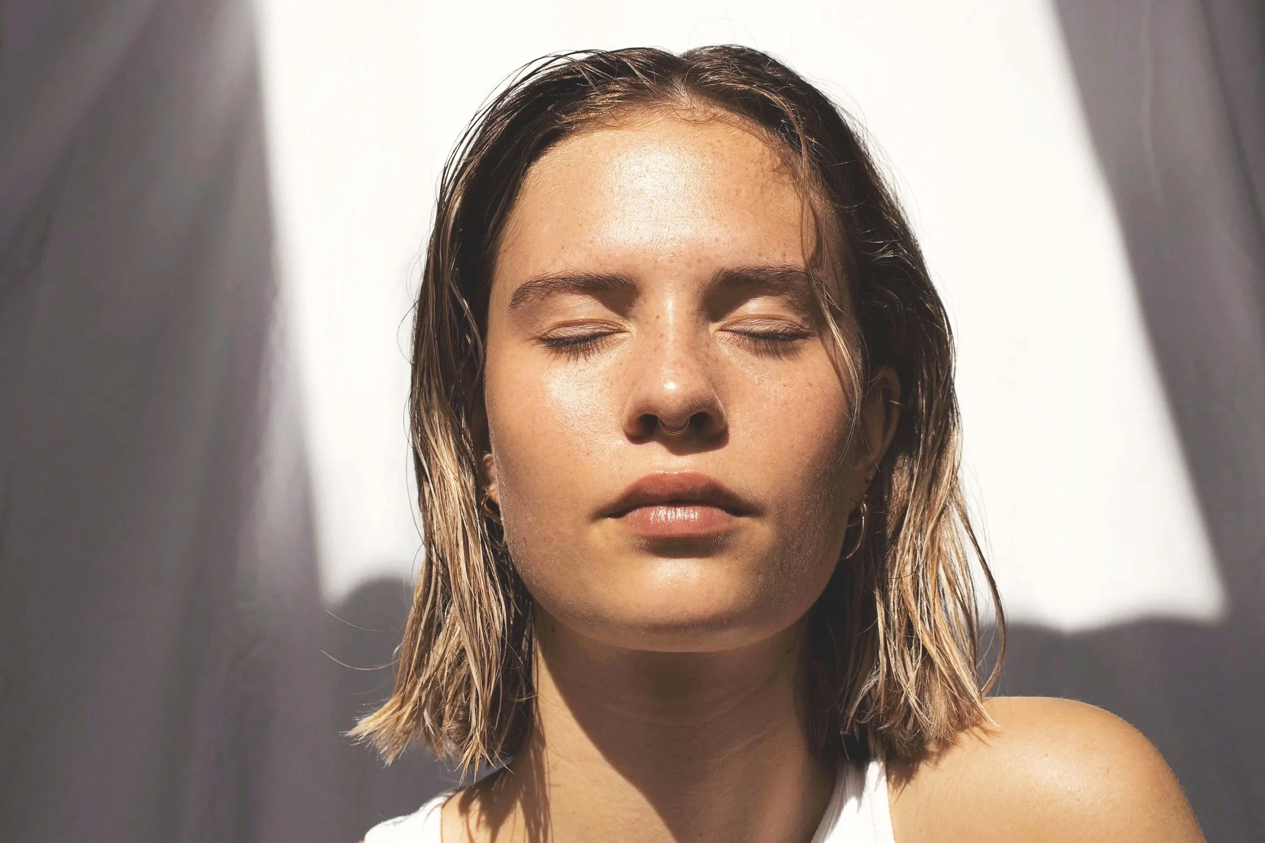 Femme aux yeux fermés, avec des cheveux mouillés, en lumière naturelle, fond blanc.