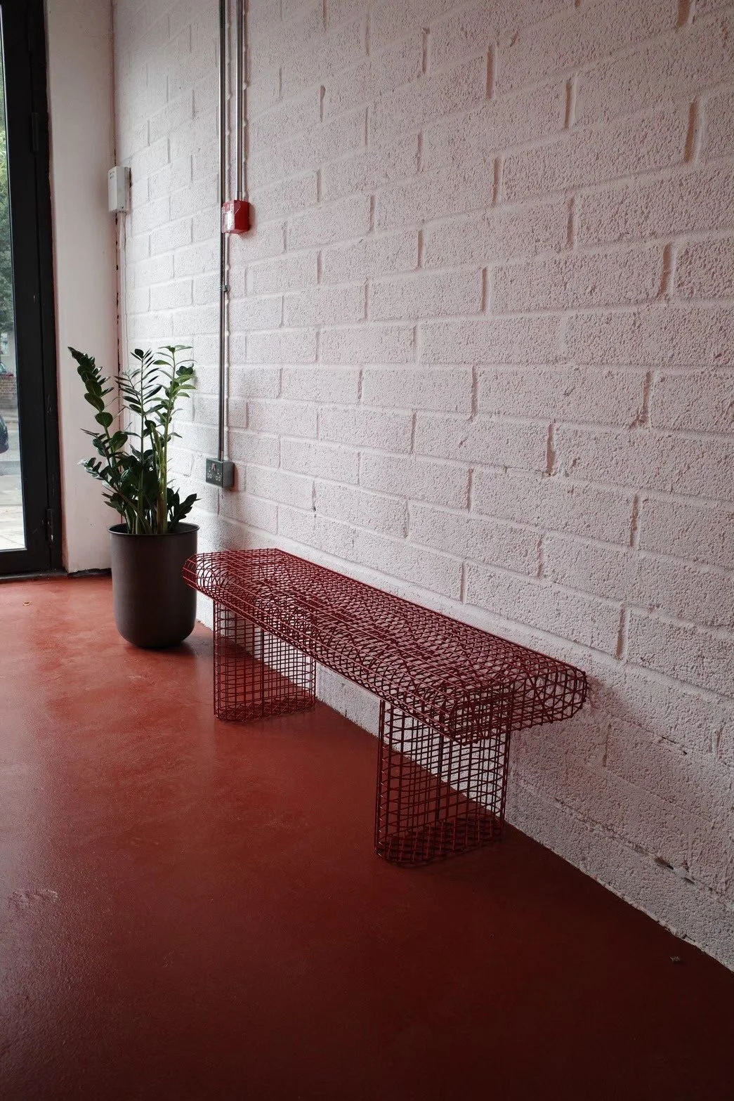 A red wireframe bench next to a large potted plant on a red floor against a white brick wall with exposed piping and a fire alarm.