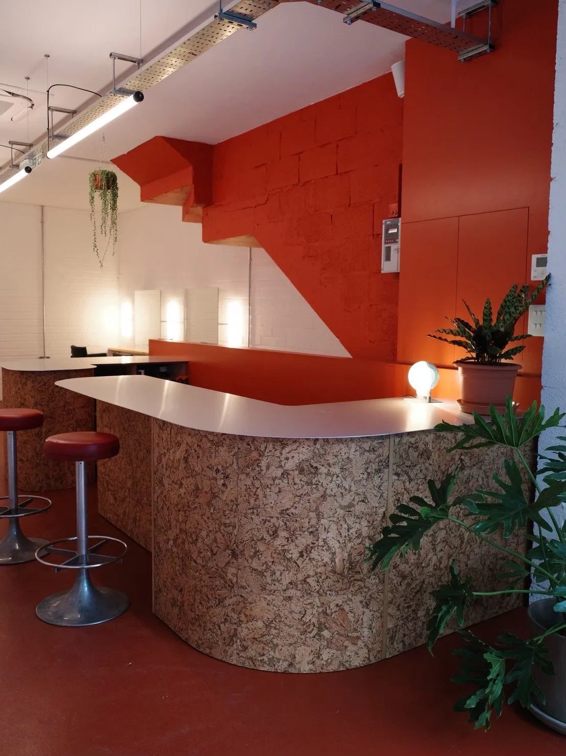 Modern interior with a curved terrazzo bar counter, green potted plants, a small spherical lamp, and orange and white walls with lighting fixtures.