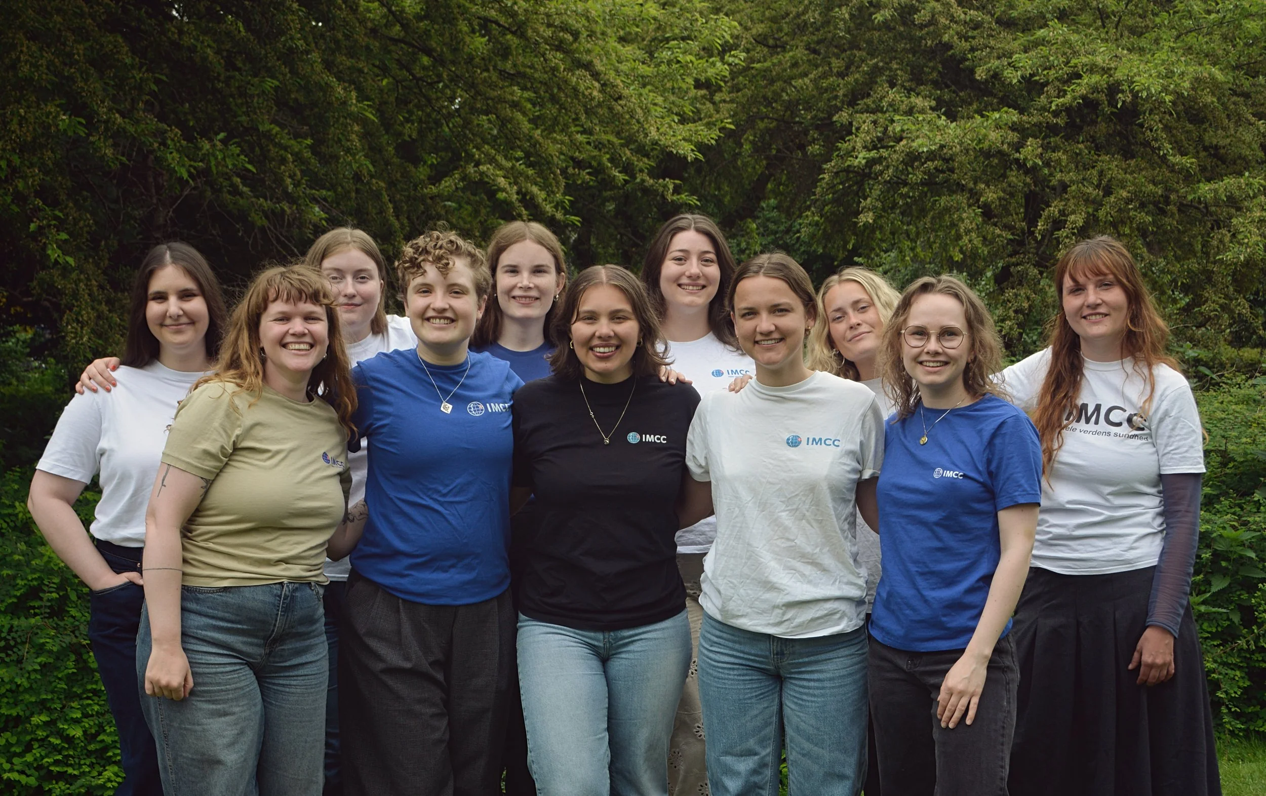 Fællesbillede af sekretariatet i grønne omgivelser. Fra venstre: Adrienn, Amanda, Kirstine, Katla, Nicoline, Eva, Freja, Julie, Kamma, Tilde og Laura. Alle iklædt T-shirts med IMCC-logoet.