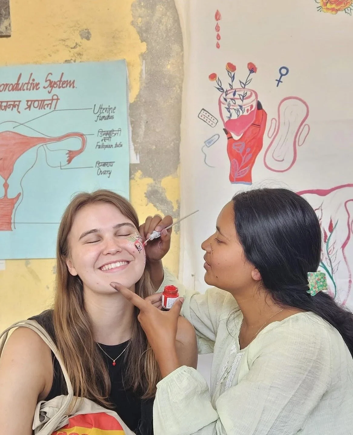 Billede fra Nepal. To kvinder, hvor den males og og den anden bruger en pensel til at lave ansigtskunst. Baggrund med anatomiske diagrams af det reproduktive system.