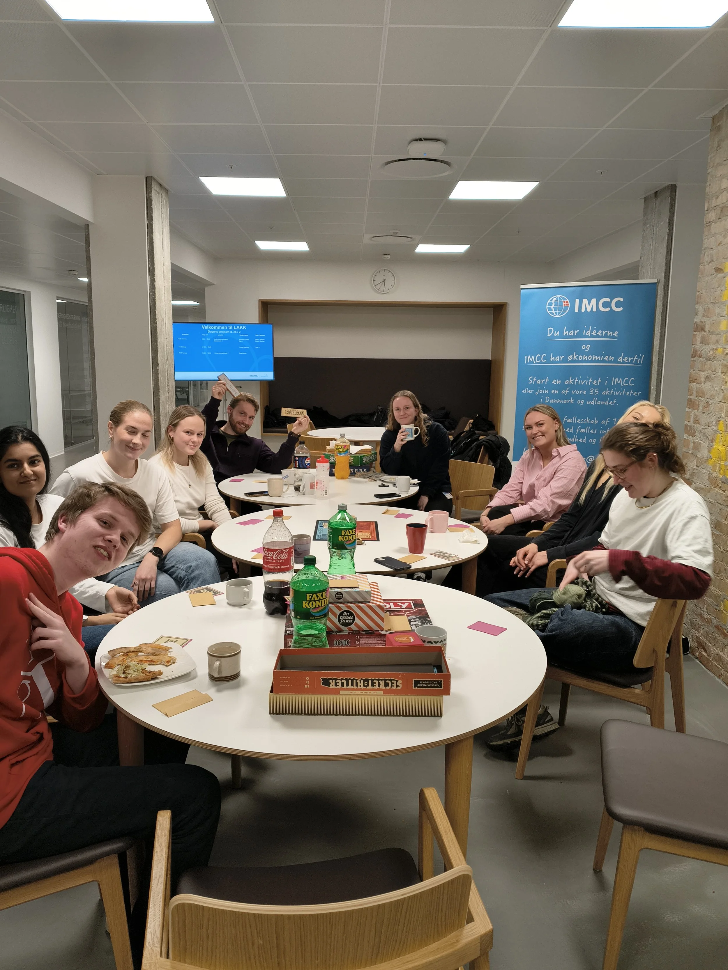 Gruppe mennesker sidder rundt om et bord og nyder mad og drikke i et konferencelokale. Der er tallerkener med pizza, sodavand, kopper og spil på bordet. Der er en blå informationsbanner til højre og en TV-skærm bagved. Møbler som stole og borde, samt en væg med en uro og en væg med en bræmme af mursten.
