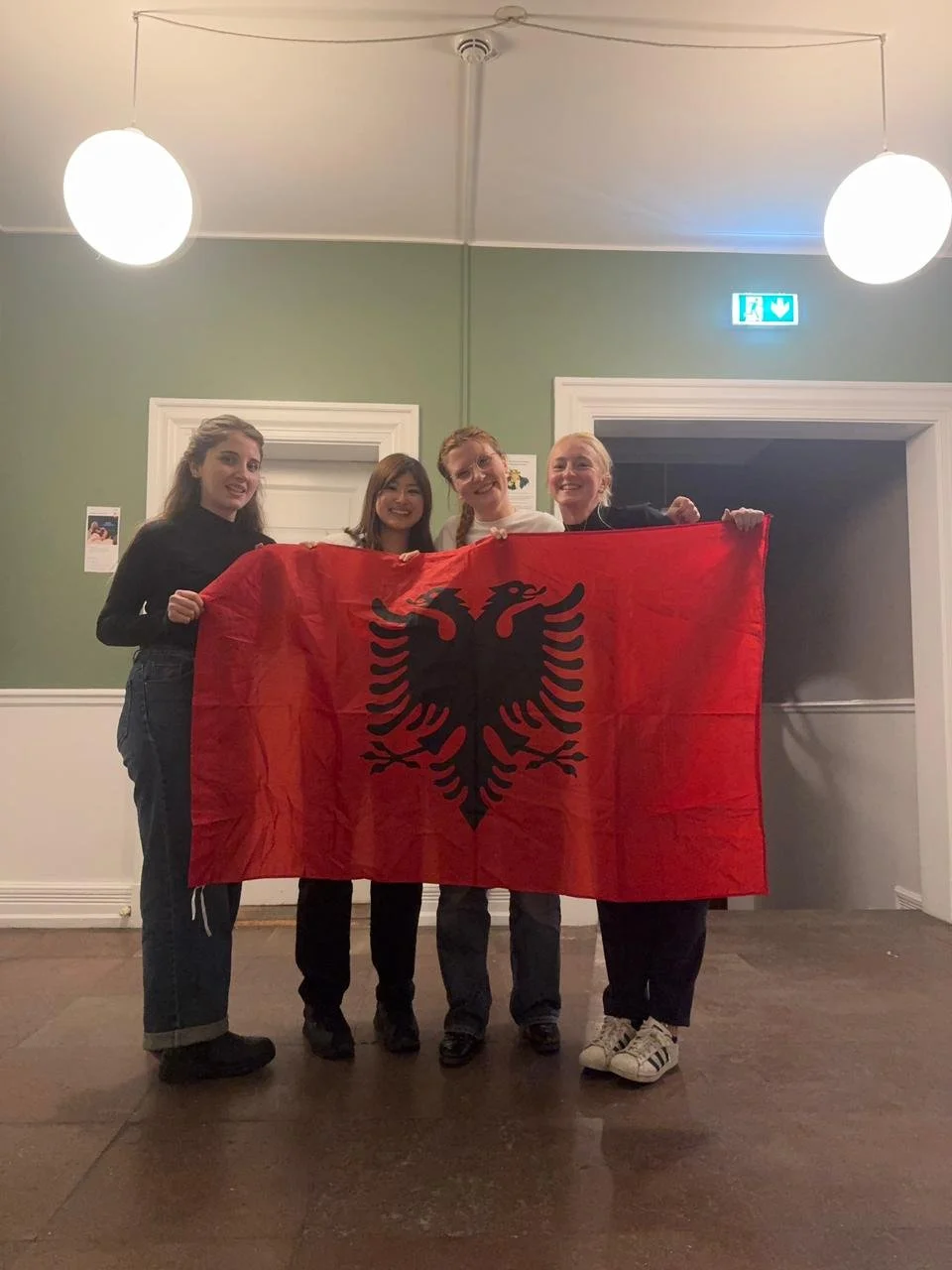Fire kvinder står sammen og holder et rødt albansk flag med en sort ørn i midten.