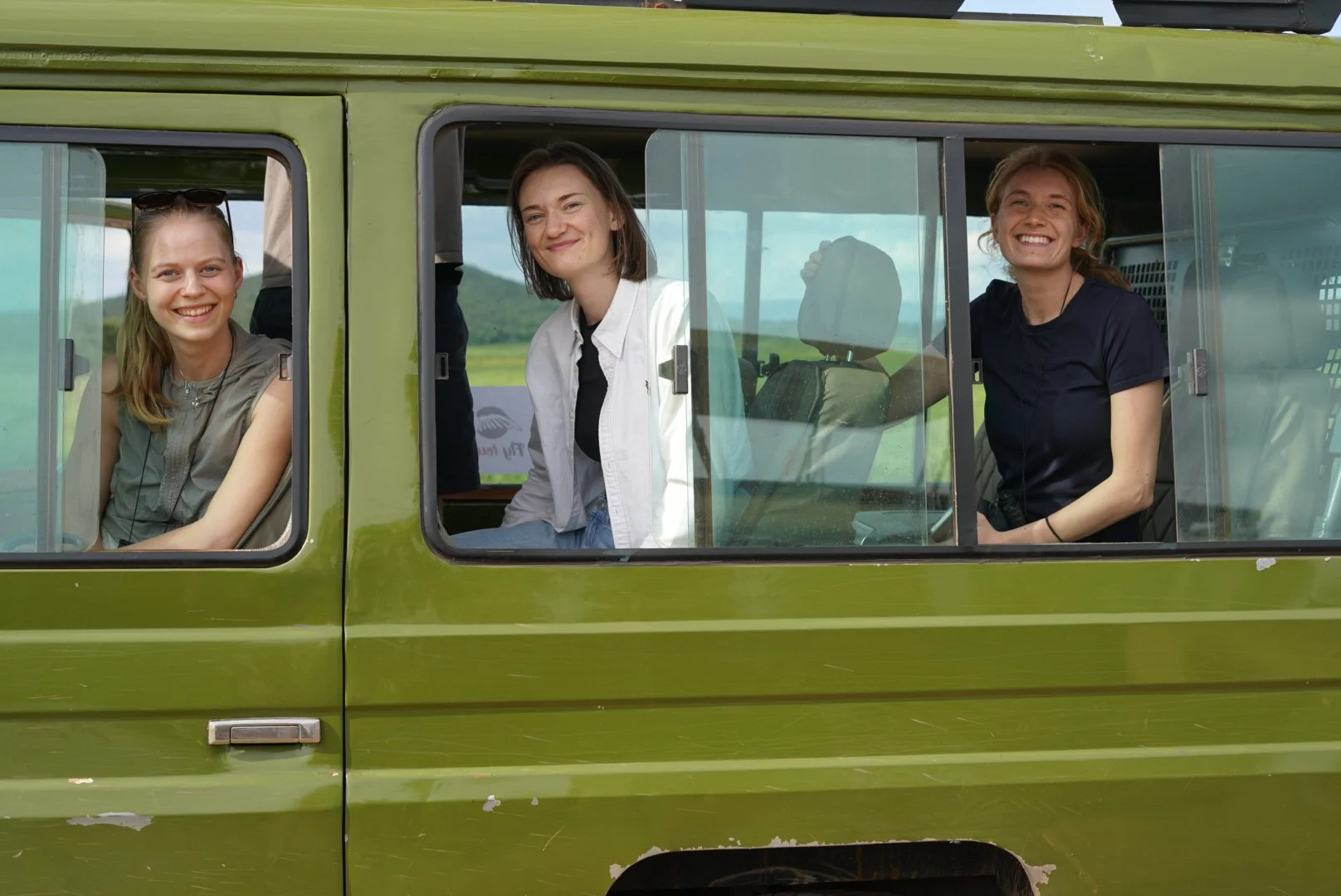 Tre kvinder sidder i en grøn bus, smilende og ser mod kameraet, i et landligt område.