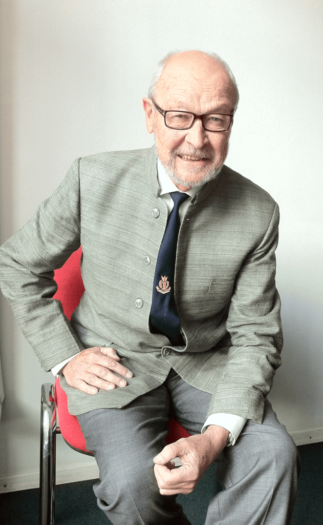 Et billede af Erik Holst, den første formand i IMCC. På billedet er han en ældre mand med briller, iført en grå jakke og en sort slips. Sidder på en rød stol og smiler.
