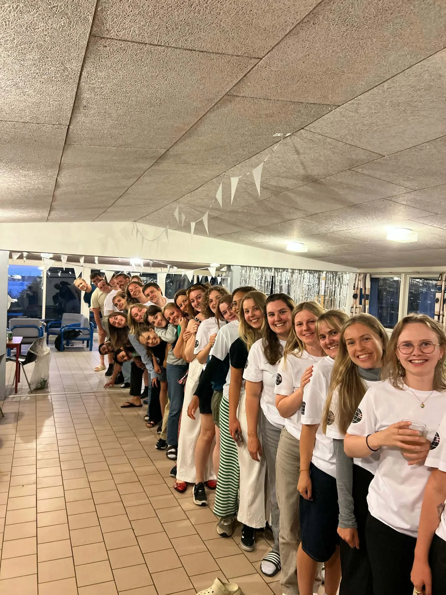 En gruppe mennesker står i en linje og smiler, hvilket tyder på en festlig sammenkomst i et indendørs miljø med store vinduer.
