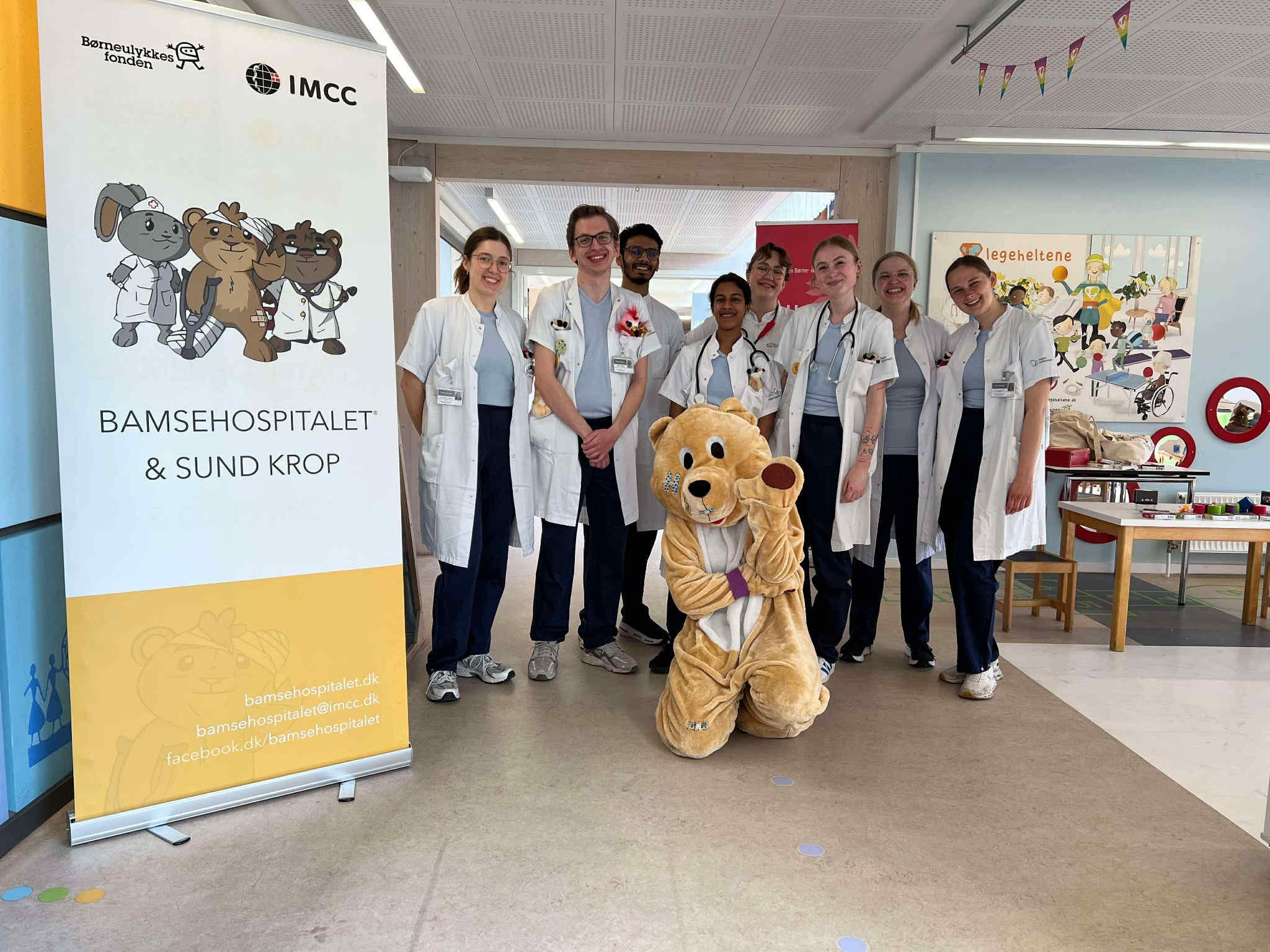 Syv personer i hospitalsskjorter står sammen foran en plakat med tegneseriehelter og en maskot i en hospitalsgang.