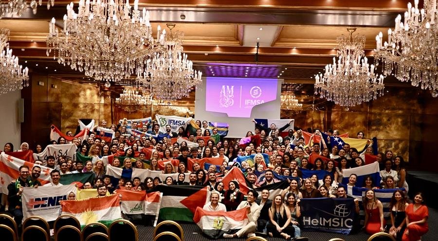 Billede fra en international konference. Gruppe mennesker i et konferencelokale, mange holder flag fra forskellige lande, foran en stor skærm med logoer og tekster.
