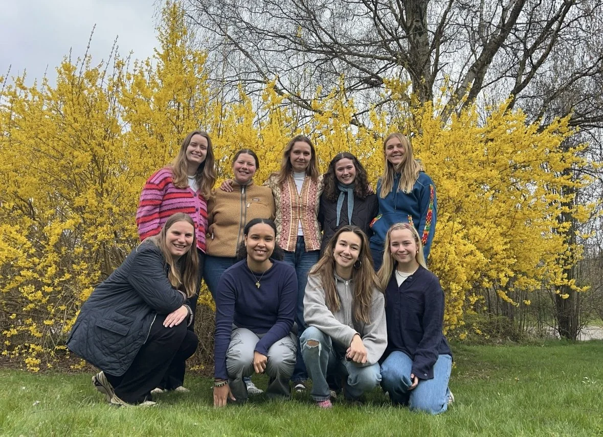Ti unge kvinder smiler foran en blomstrende gyldenbusk i en park.