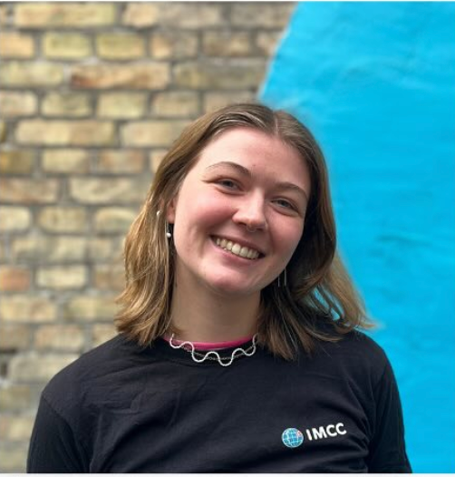 En smilende kvinde med brunt hår, iført en sort T-shirt med IMCC-logo, står foran en murstensvæg og en blå struktur.