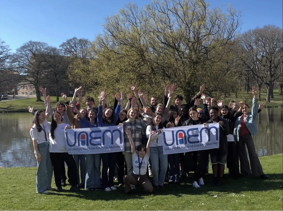 Gruppe unge mennesker holder op foran en dug med teksten 'UAEM' og 'Universities Allied for Essential Medicines', ved en sø i en park med træer og grønne græsplæner.