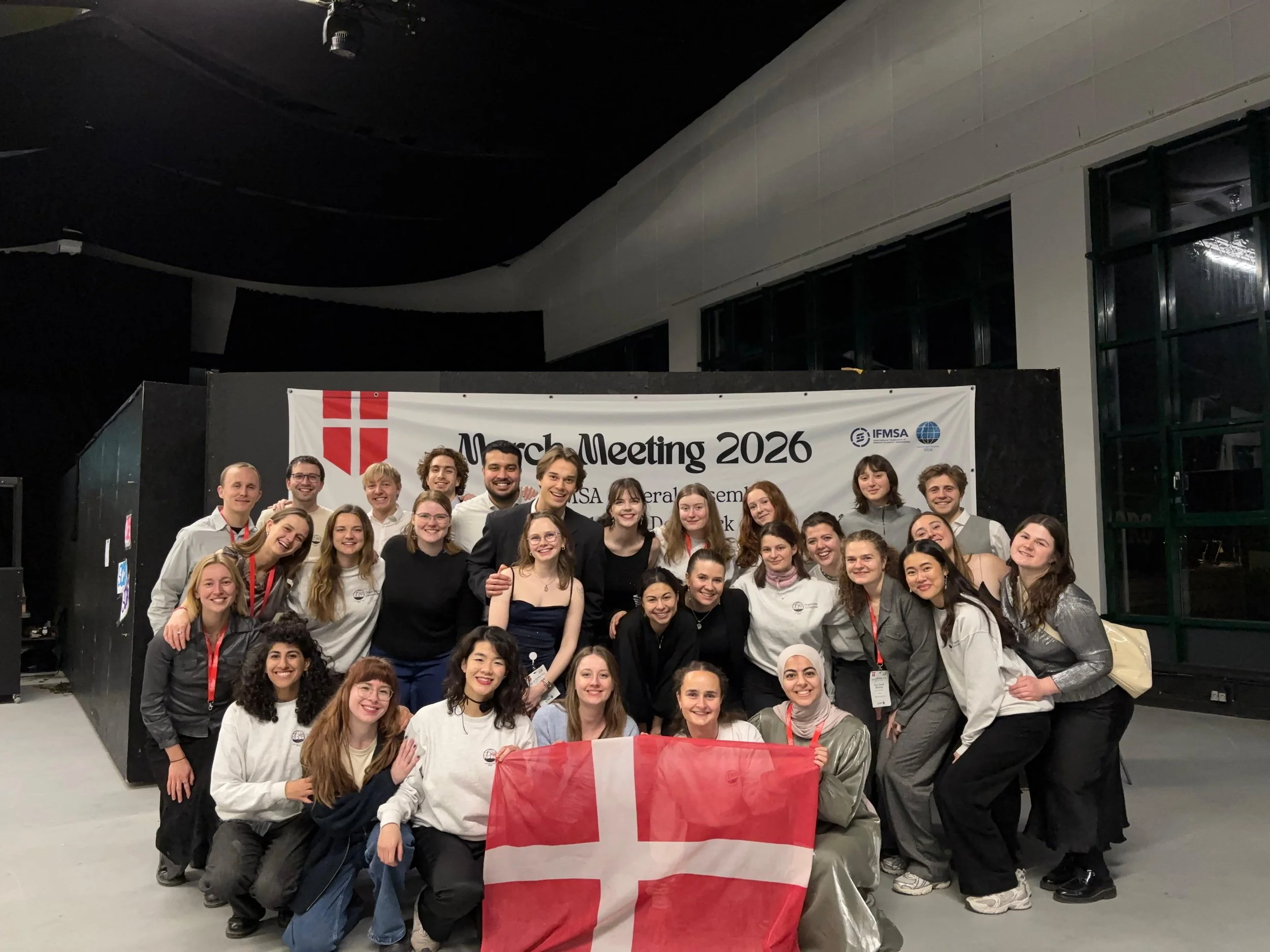 Billede fra international konference. Stort gruppe af unge mennesker foran en banner med teksten 'March Meeting 2026' og et dansk flag.