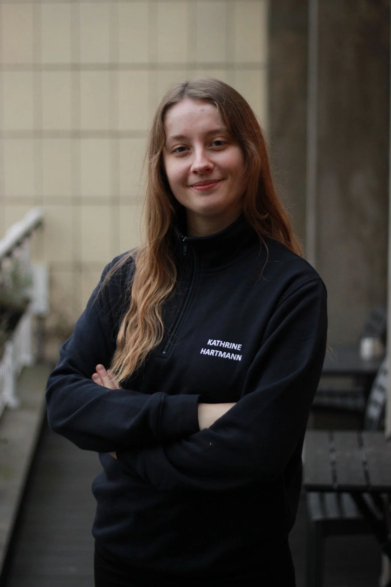Kvinde med langt, lyst hår, iført sort jakke med navn 'Kathrine Hartmann' står med armene krydset foran en busk og trapper.
