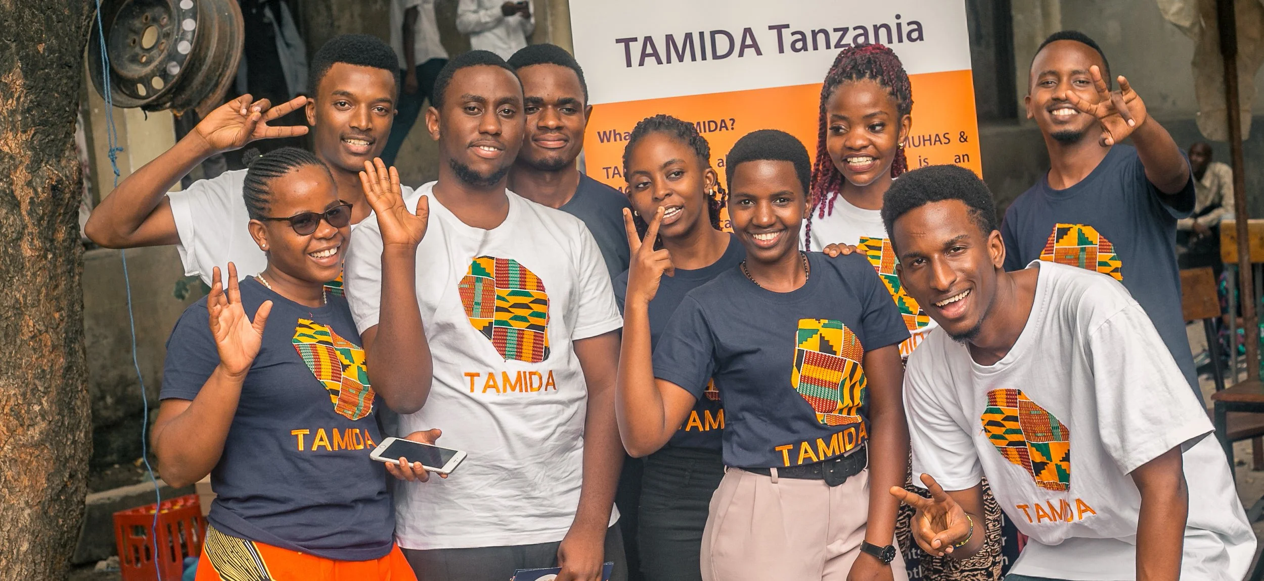 En gruppe unge mennesker, der smiler og laver fredstegn foran en Tajida Tanzania banner, alle iført T-shirts med afrikanske mønstre.