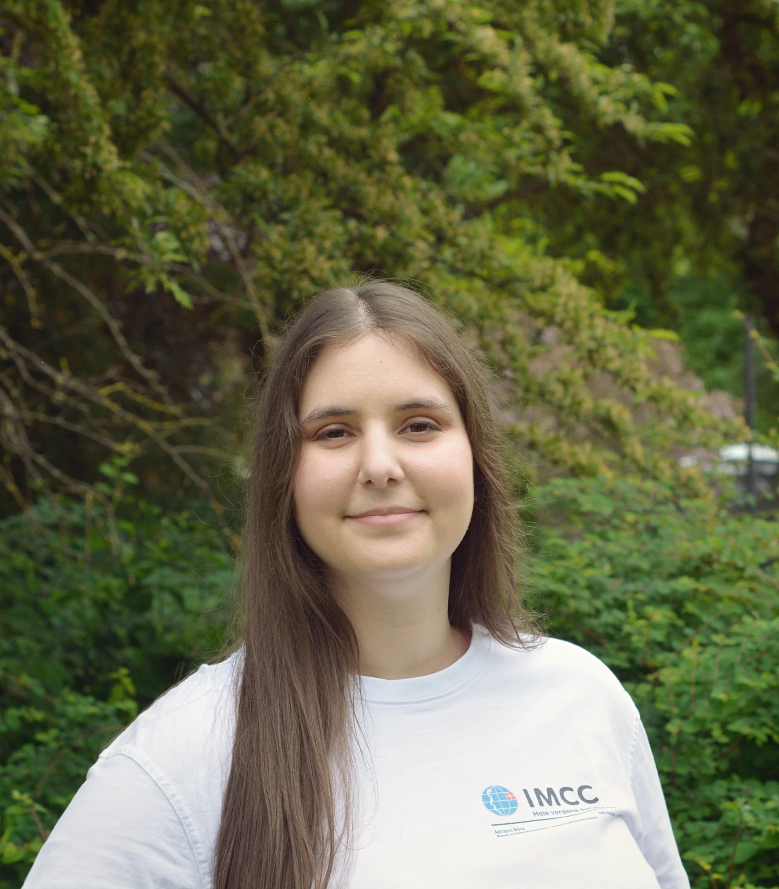Portræt af Adrienn. Langt brunt hår, smilende, iført hvid IMCC-trøje.