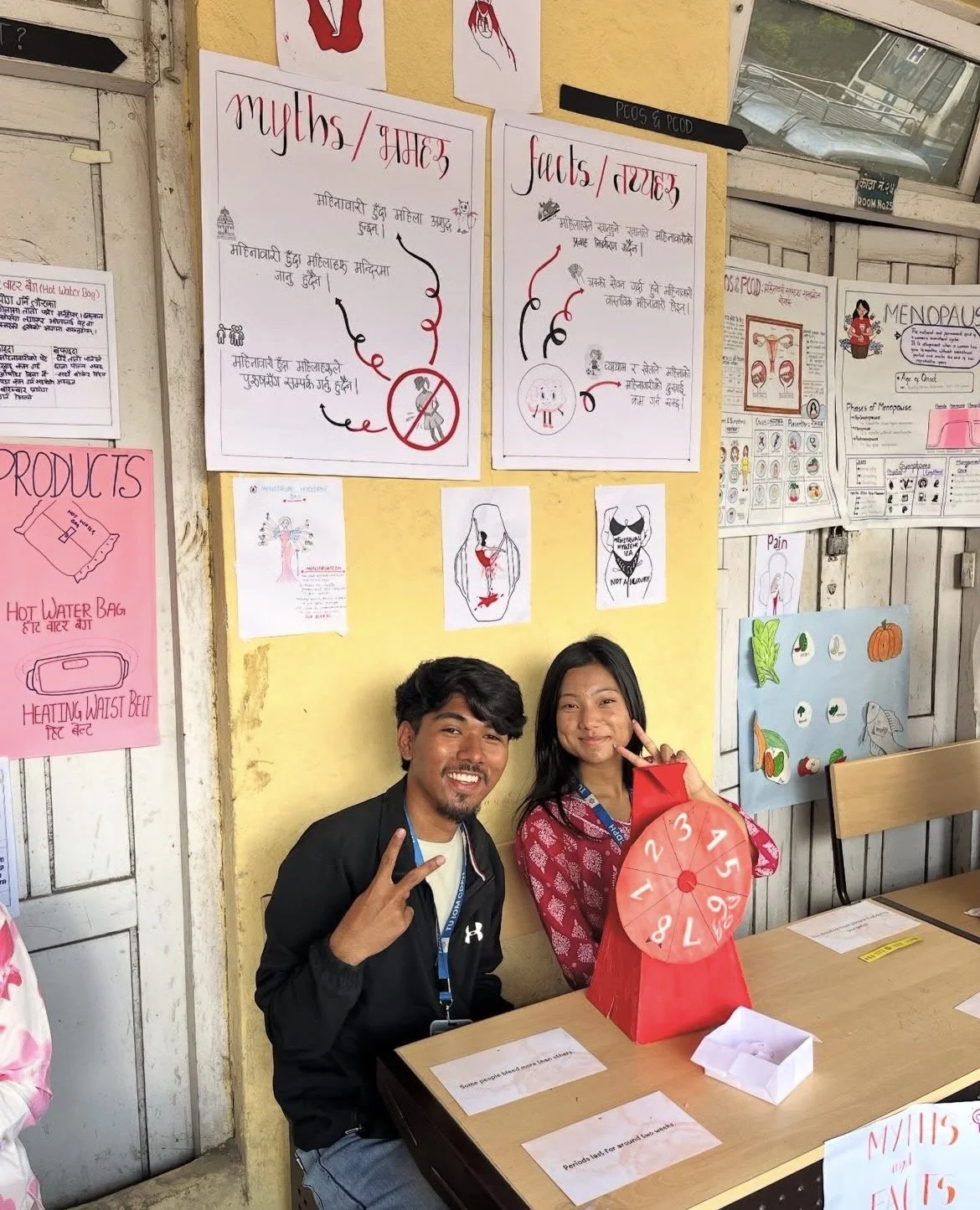 Billede fra Nepal. To personer sidder ved et bord i et klasseværelse, begge smiler og laver fredstegn med hænderne. På væggen bag dem er der flere informations- og uddannelsesposters.