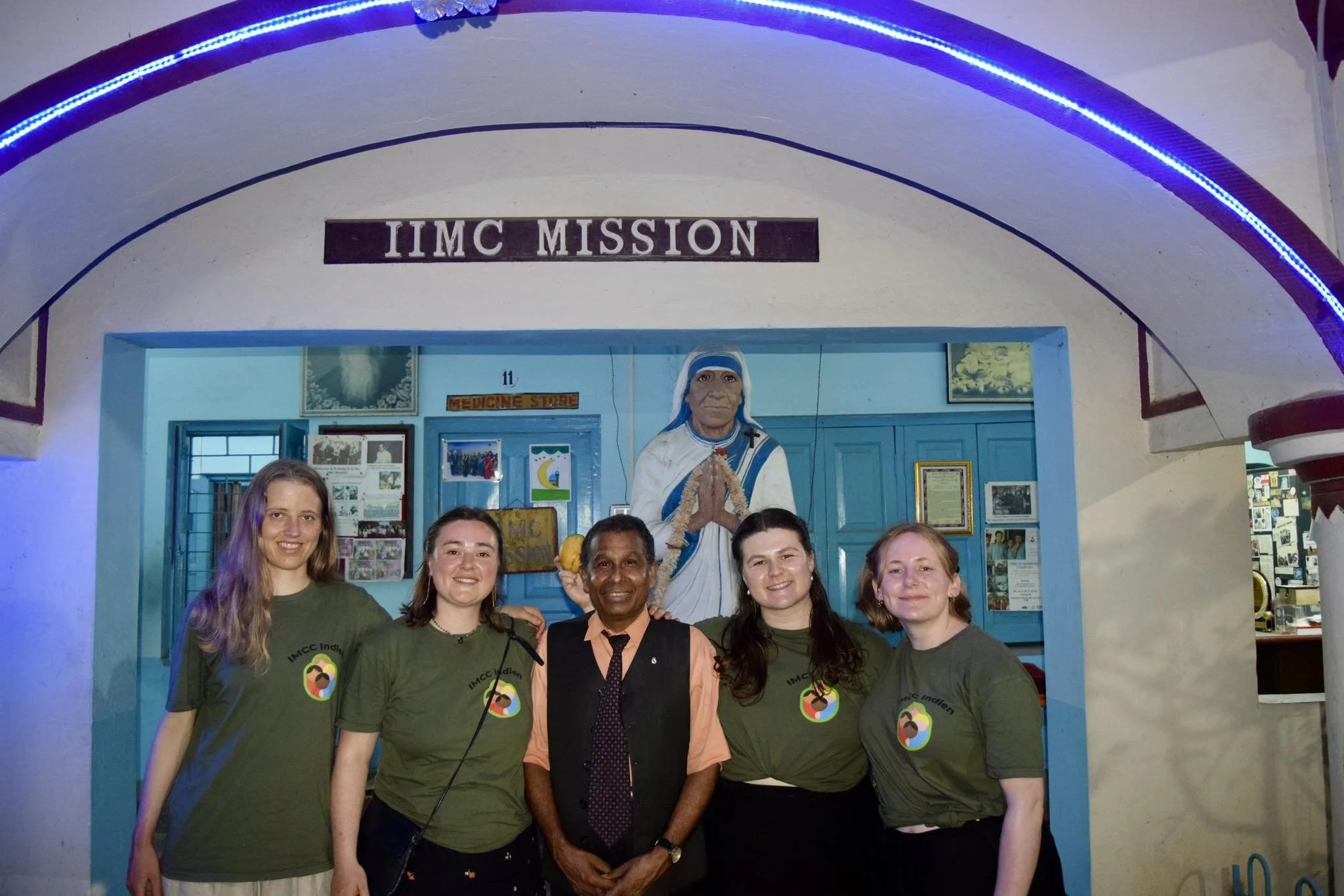 Gruppe af fem mennesker står foran en blå dør med en statue af Jomfru Maria i baggrunden, på en bygning med skiltet 'IIMC MISSION'.