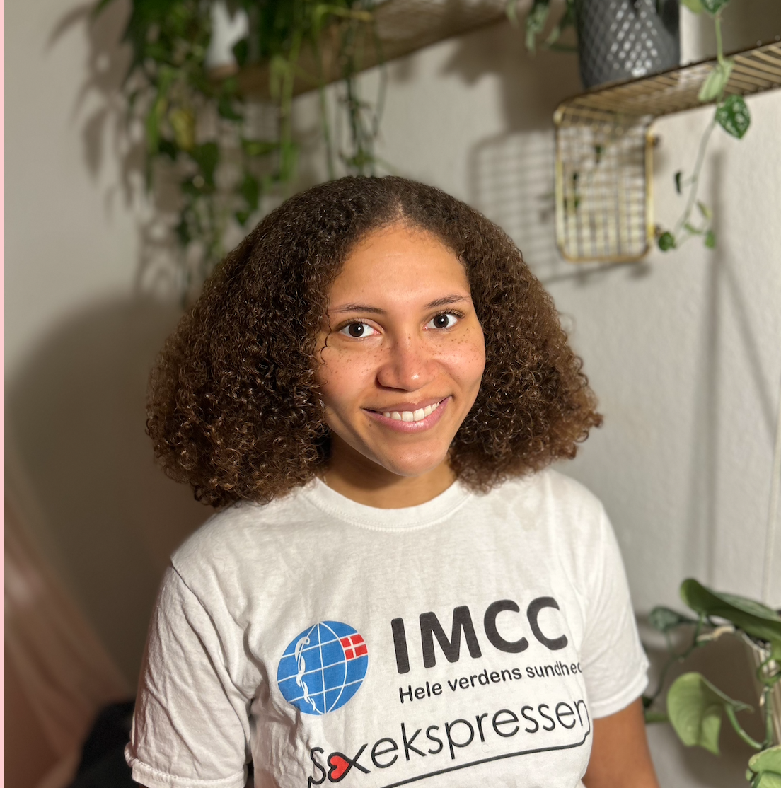 Kvindelig person med krøllet brunt hår, smilende, iført hvid T-shirt med tekst om IMCC og Verdens sundhed.