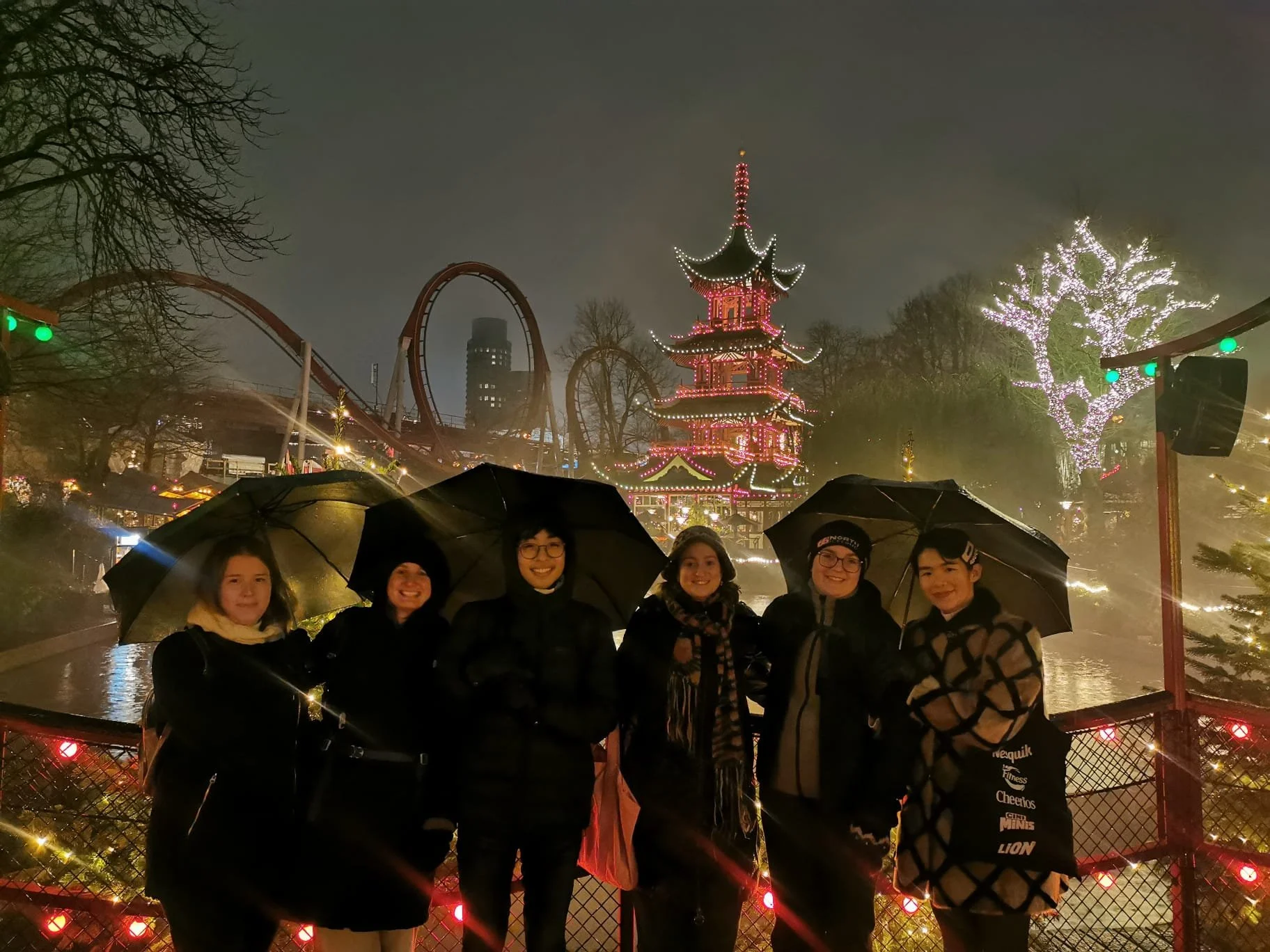 Fem personer står i regn med paraplyer foran en lystop på en forlystelsespark om aftenen, med julelys og et tempeltag i baggrunden.