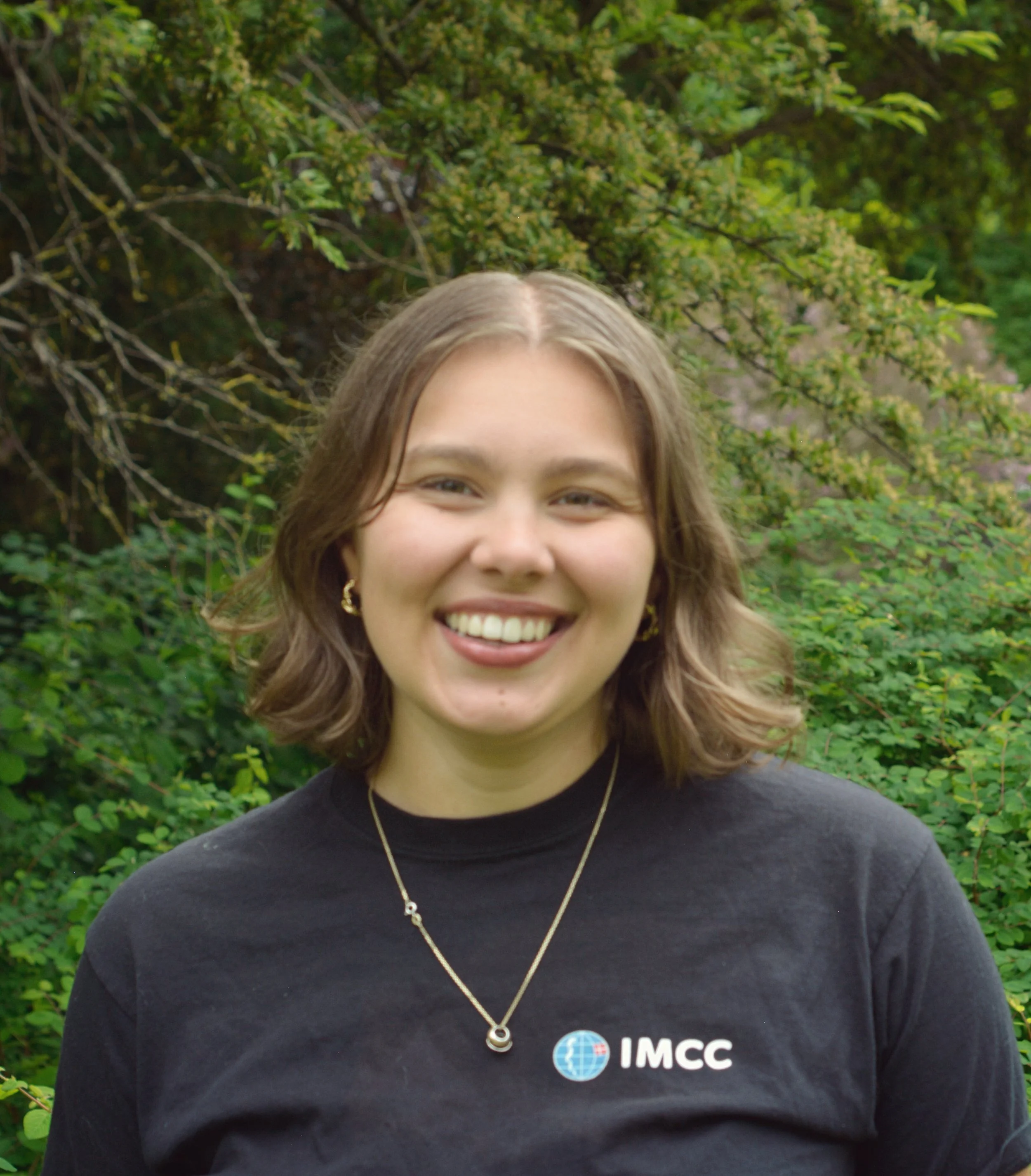 Portræt af Eva. Skulderlangt brunt hår, smilende, iført sort IMCC-trøje.