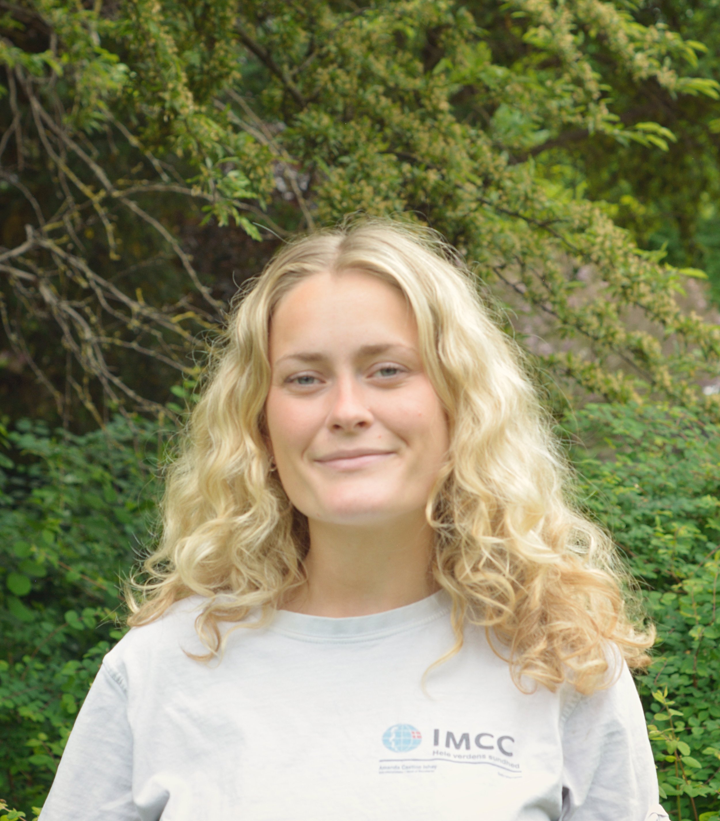Portræt af Kamma. Langt blonde krøllet hår, smilende, iført hvid IMCC-trøje.