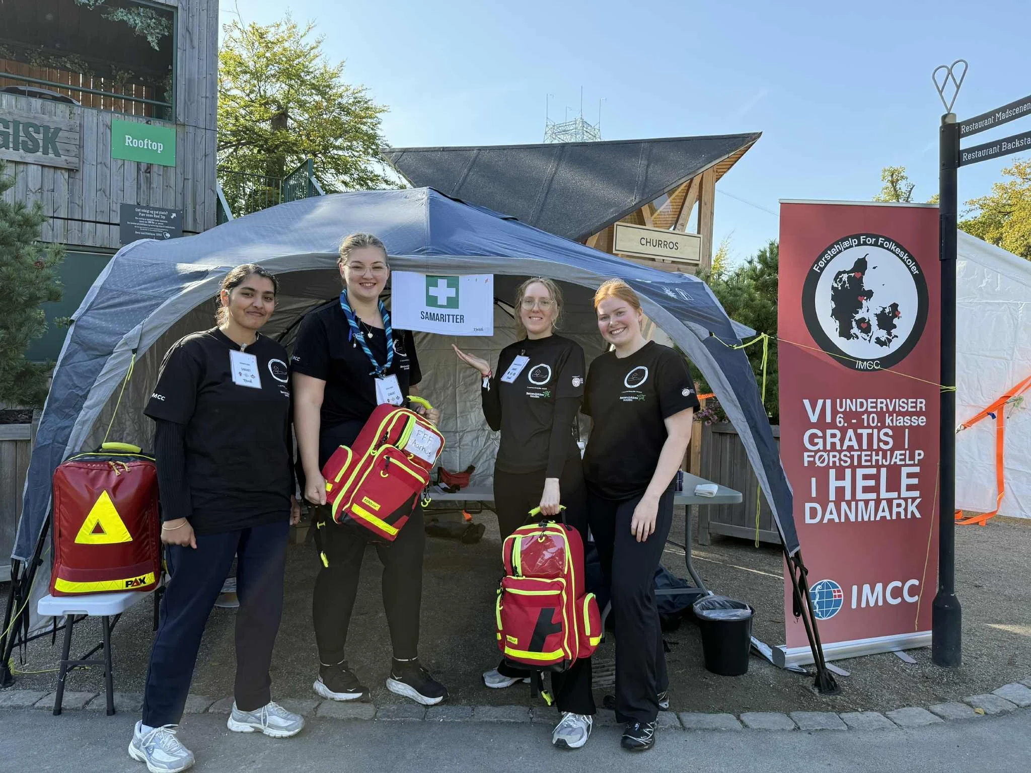 Fire unge personer iført sorte uniformer med ambulanceudstyr, der står foran en telt med en skilt, der siger "Samartitter". Der er også et banner med teksten "Vi underviser 6.-10. klasse gratis i førstehjælp i hele Danmark."