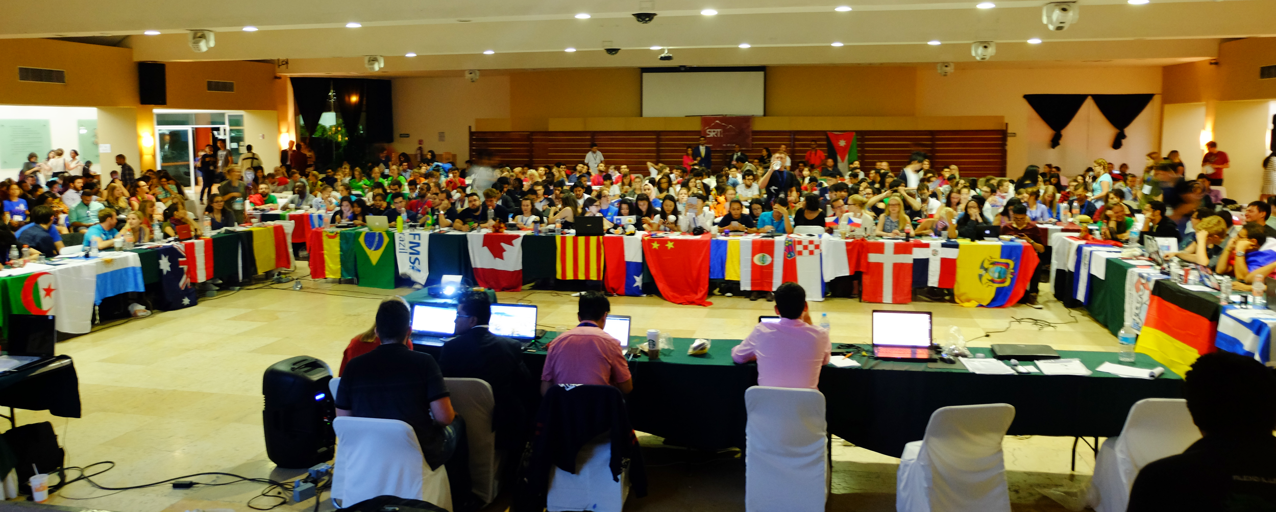 Billede fra international konference. Billede af et stort konferencelokale fyldt med mennesker, der sidder ved borde dækket med flag fra forskellige lande.