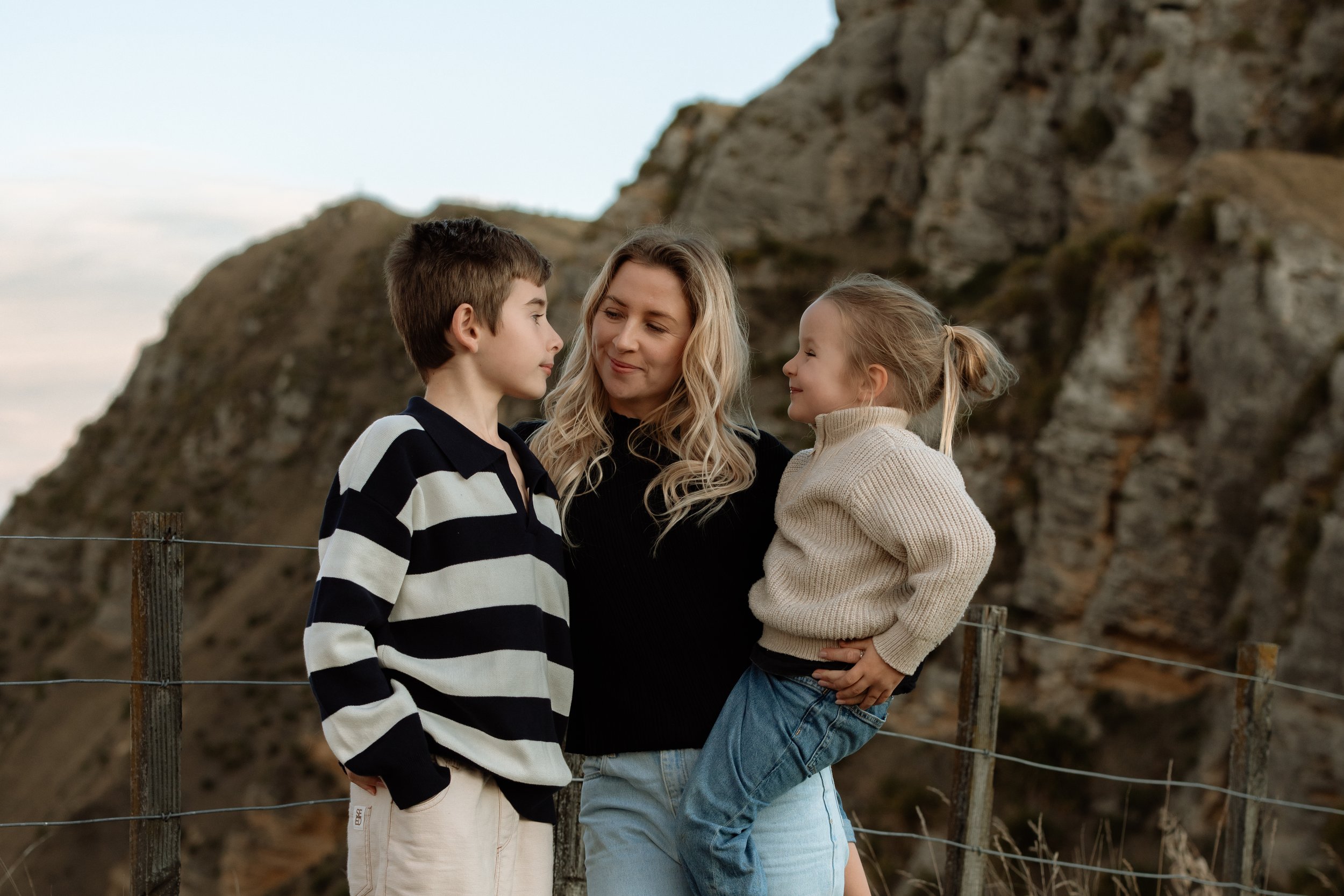 A woman with blonde hair standing between a young boy and girl outdoors near rocky cliffs, all smiling and looking at each other, dressed casually, in front of a wooden fence.