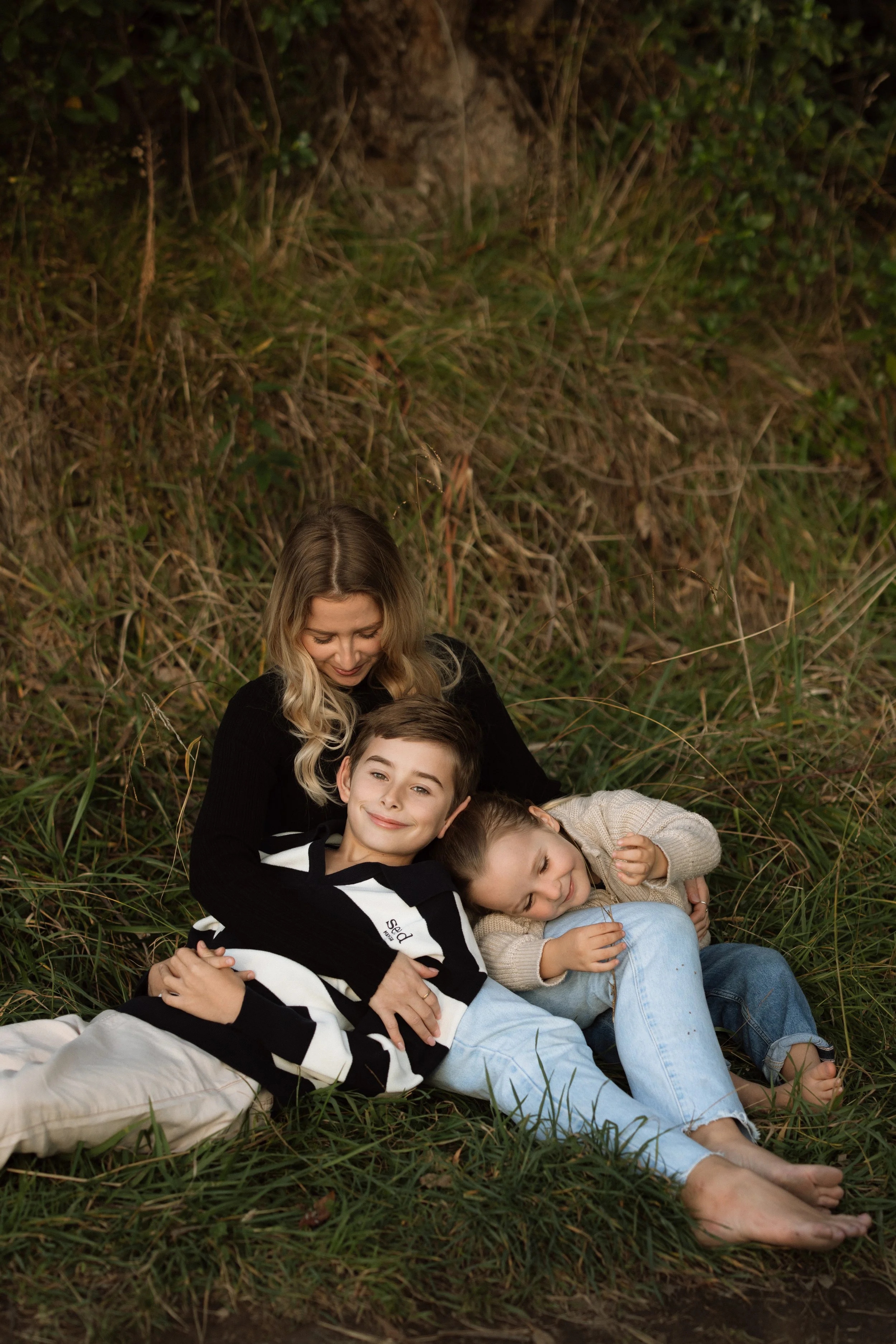 A family of three lying on the grass and leaning against each other, surrounded by tall grass and bushes, enjoying time together outdoors.