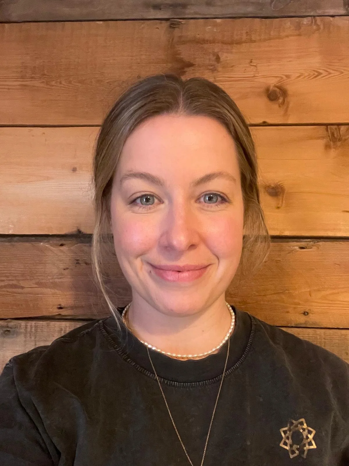 A female massage therapist with light skin, blue eyes, and brown hair smiling at the camera, wearing a black shirt with a star-shaped badge, a pearl necklace, and a gold necklace, standing against a wooden wall background.