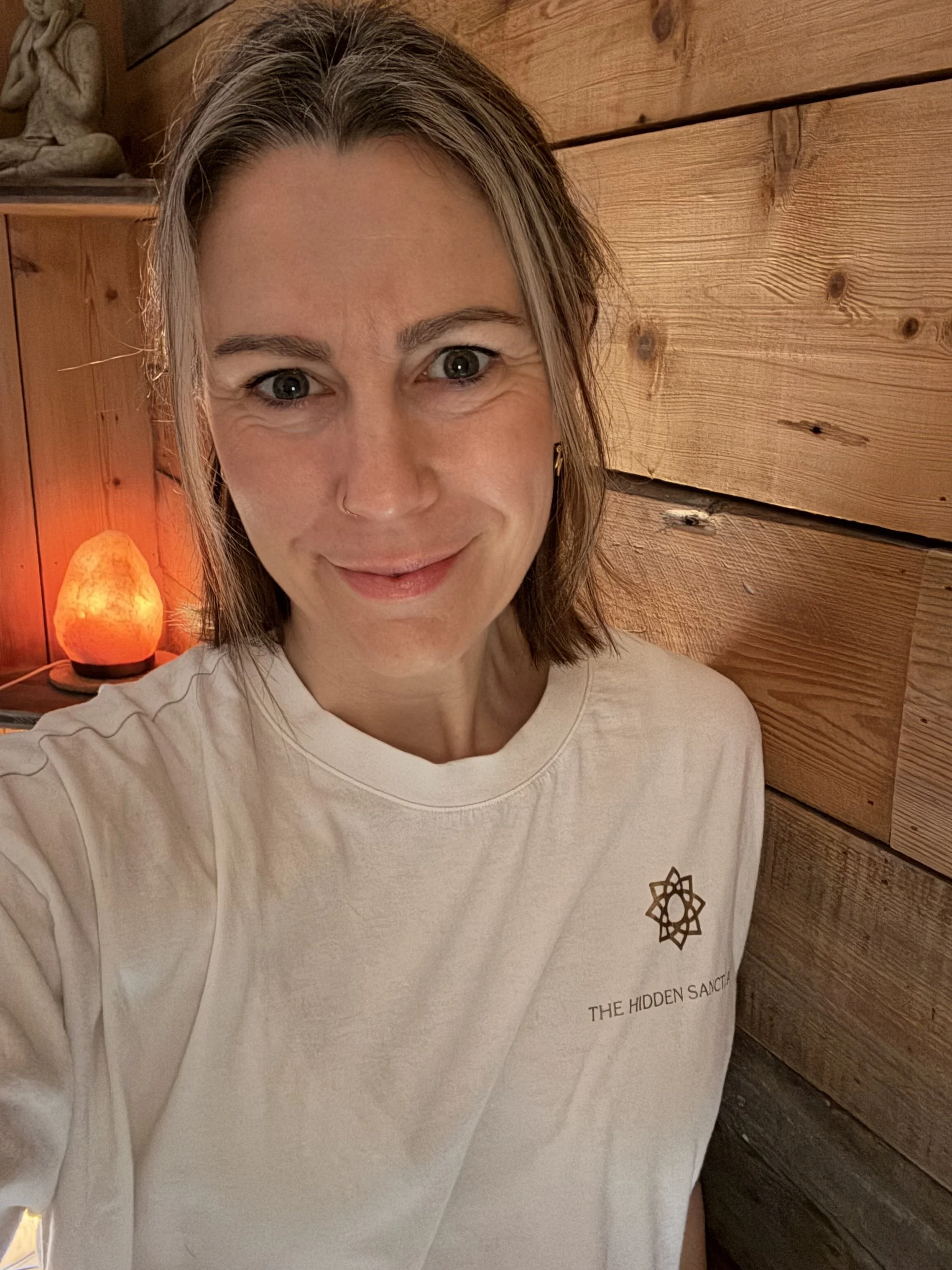 The Master Therapist with light brown hair, wearing a white T-shirt that says 'The Hidden Sanctuary' and the logo underneath,  in front of a wooden wall with a salt lamp in the background.