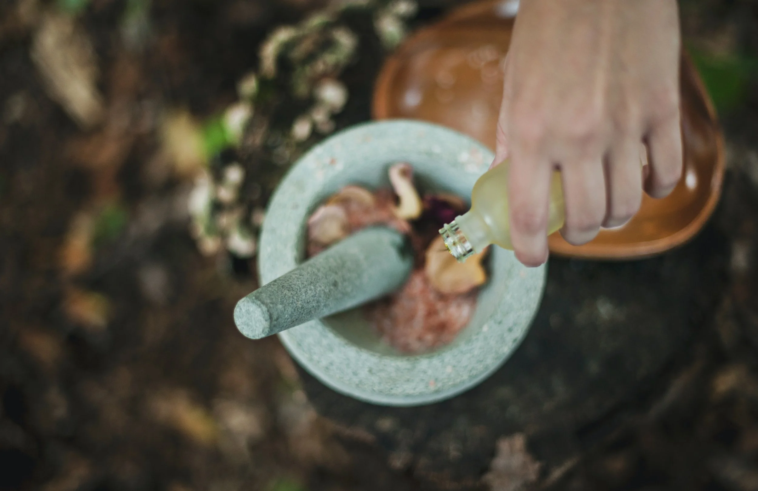 A person using a mortar and pestle to grind herbs or spices in an outdoor setting, with a hand pouring liquid from a small bottle.