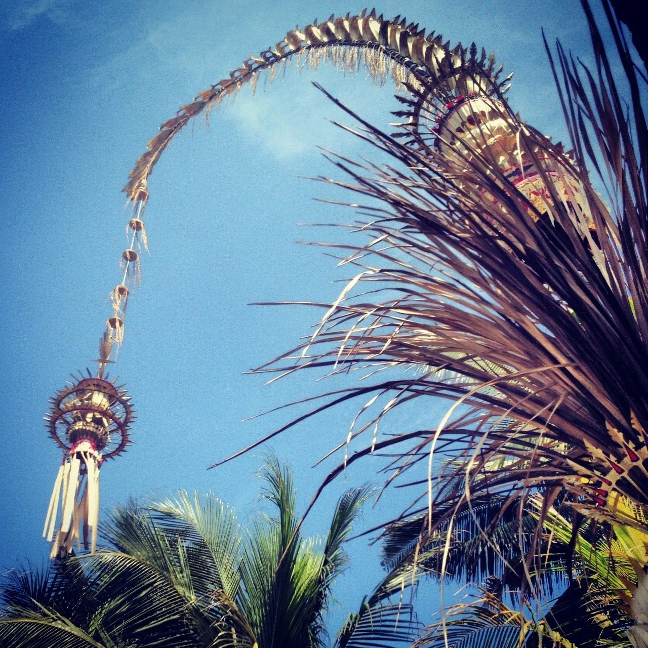 A tall decorative tower with intricate details is seen among green palm trees against a clear blue sky.