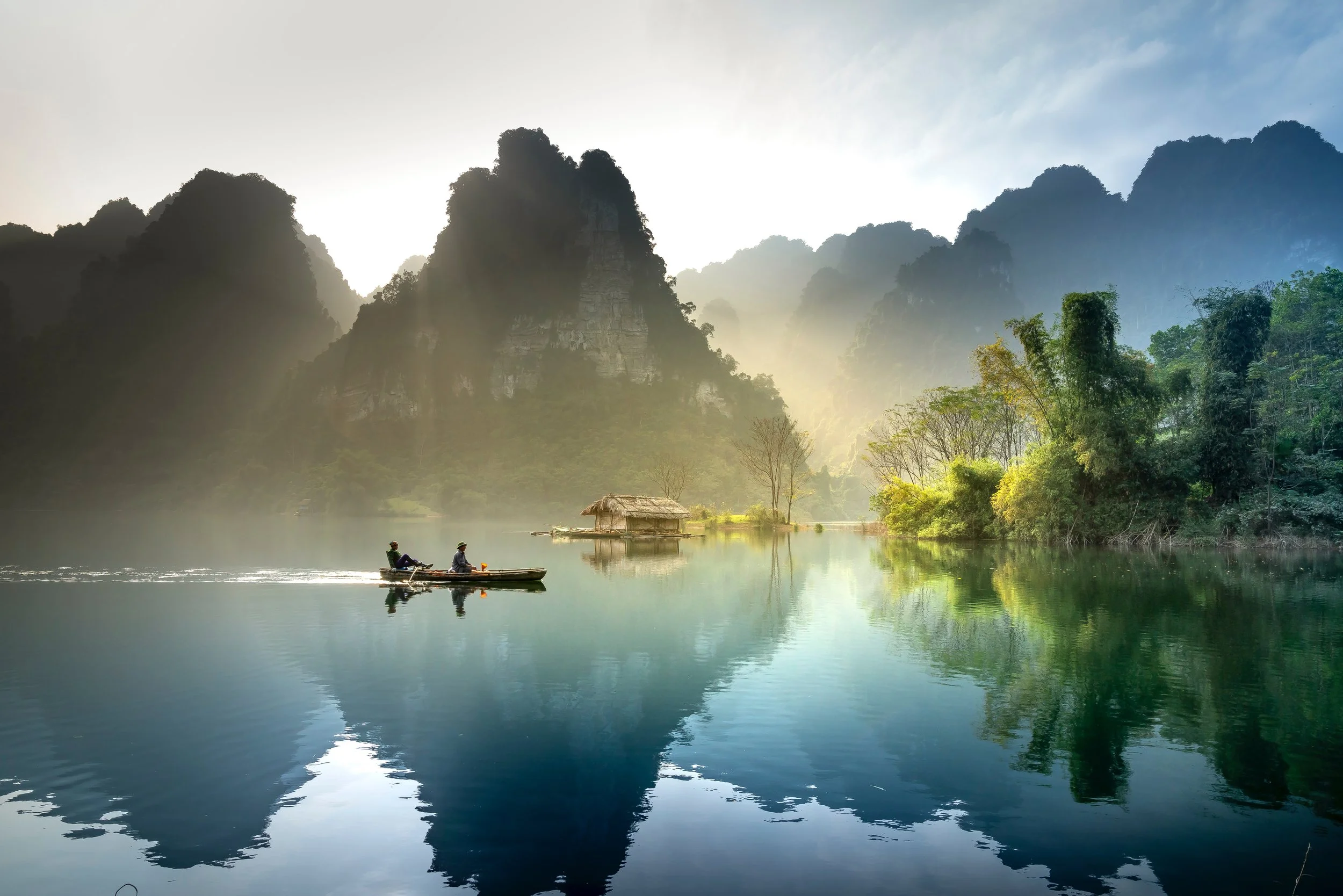 Two people riding a boat on a calm river surrounded by lush greenery and towering karst mountains, with a small hut on the riverbank and mist at sunrise or sunset.