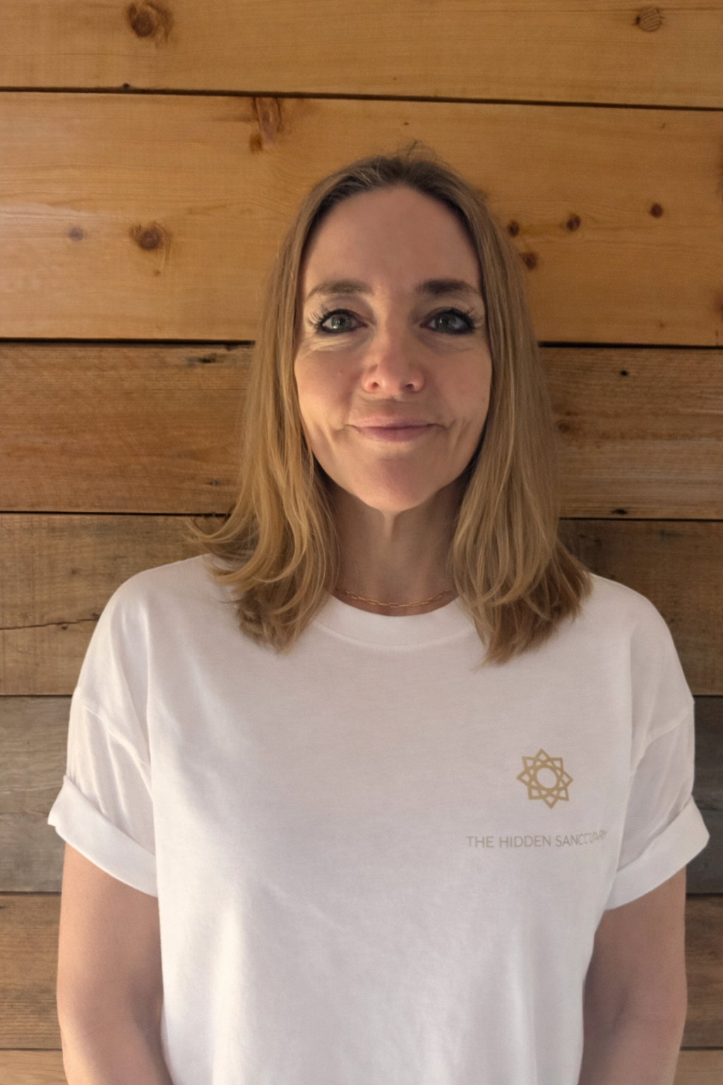A female massage therapist with shoulder-length blonde hair wearing a white t-shirt with a logo and text, standing in front of a wooden wall.